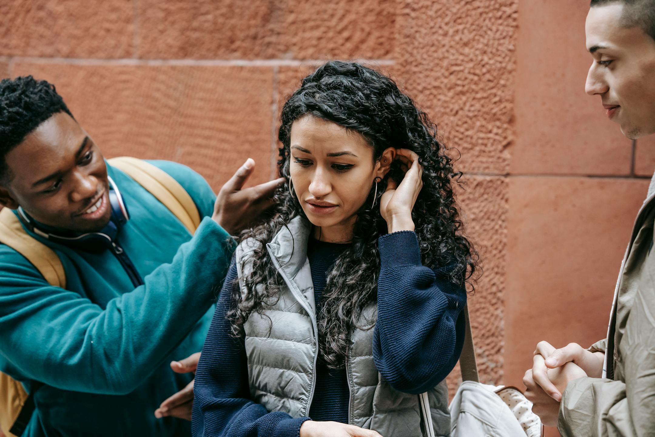 Crop African American teenage man abusing and touching hair of depressed ethnic female groupmate while standing on street with male friend