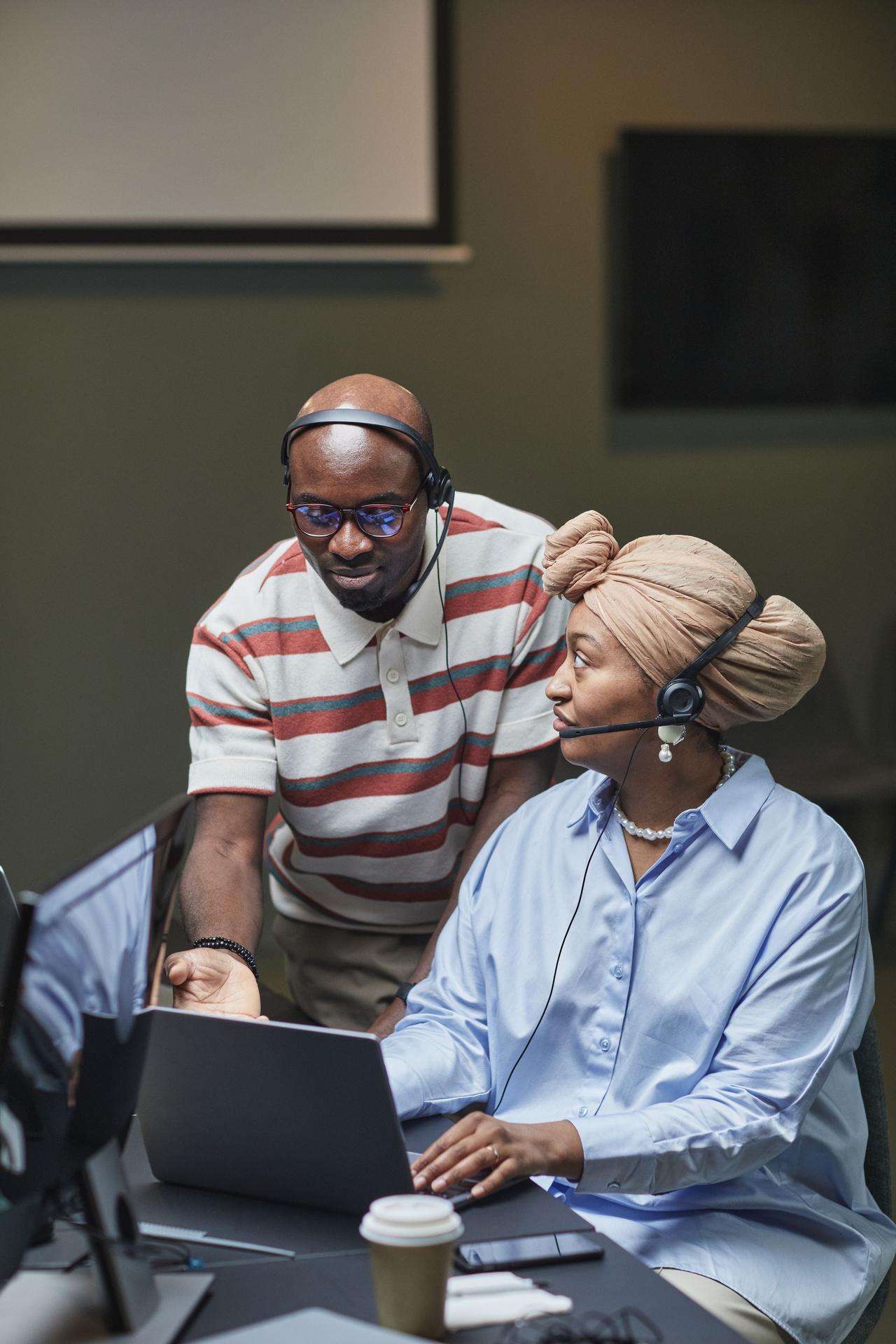 Two coworkers using laptops with headsets in an office space, engaged in a collaborative task.
