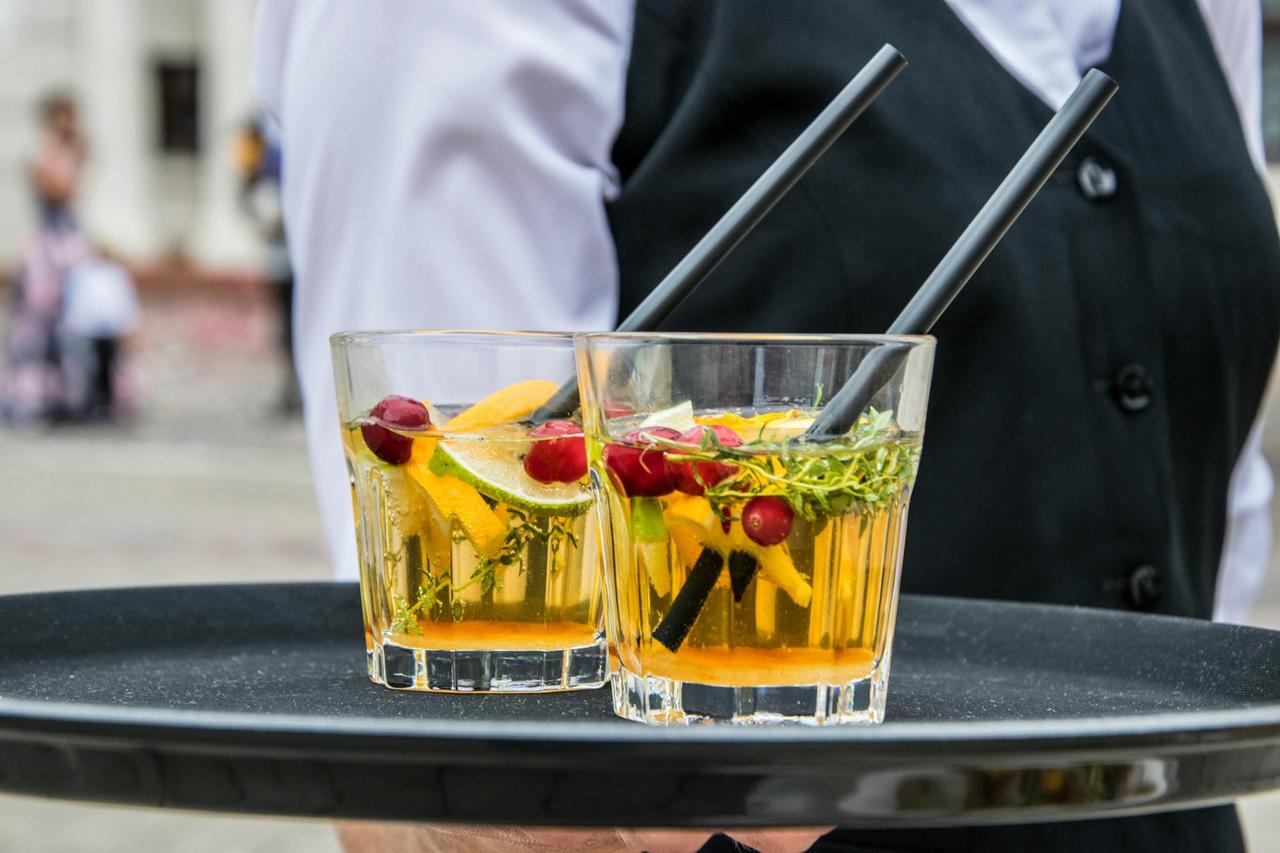 Delicious orange cocktails with cranberries and herbs on a tray, served by waiter at an outdoor event.