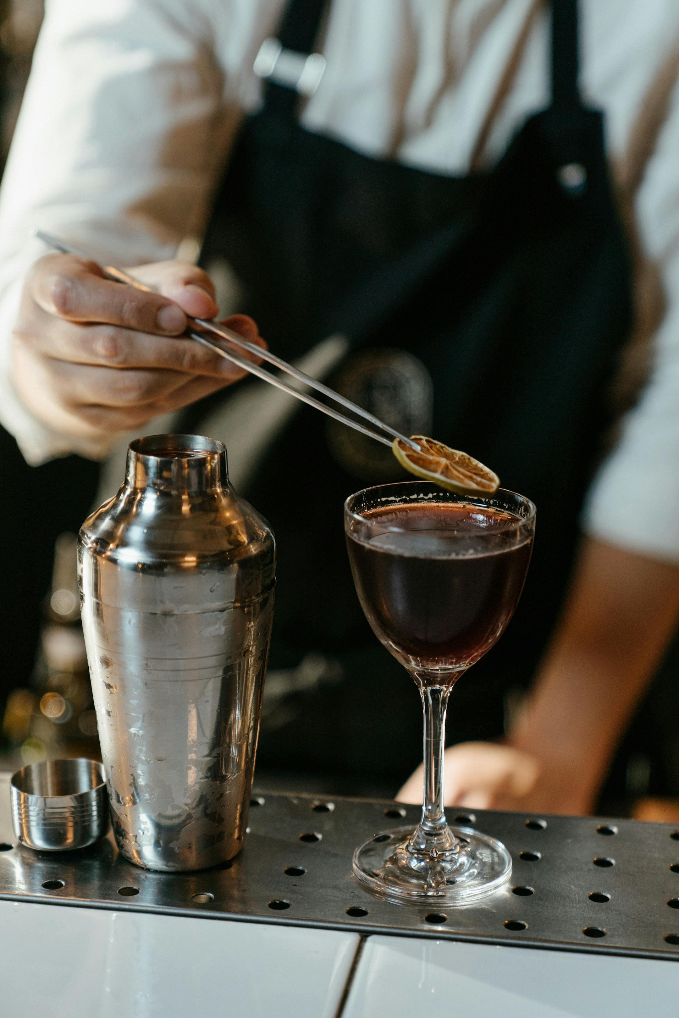 A bartender carefully adds a garnish to a cocktail using tweezers at a bar counter.