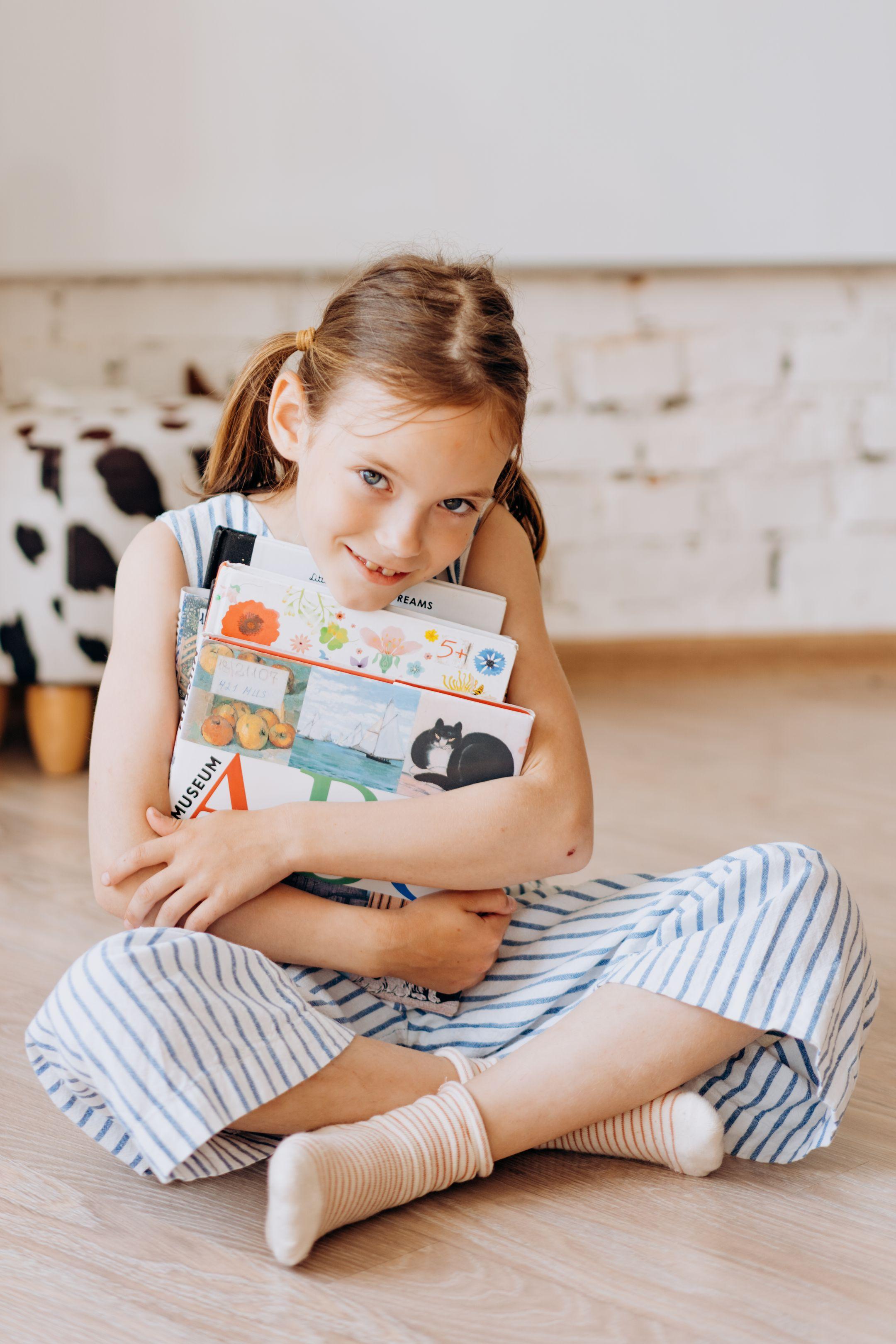 Adorable young girl hugging a stack of picture books while sitting on a wooden floor indoors.