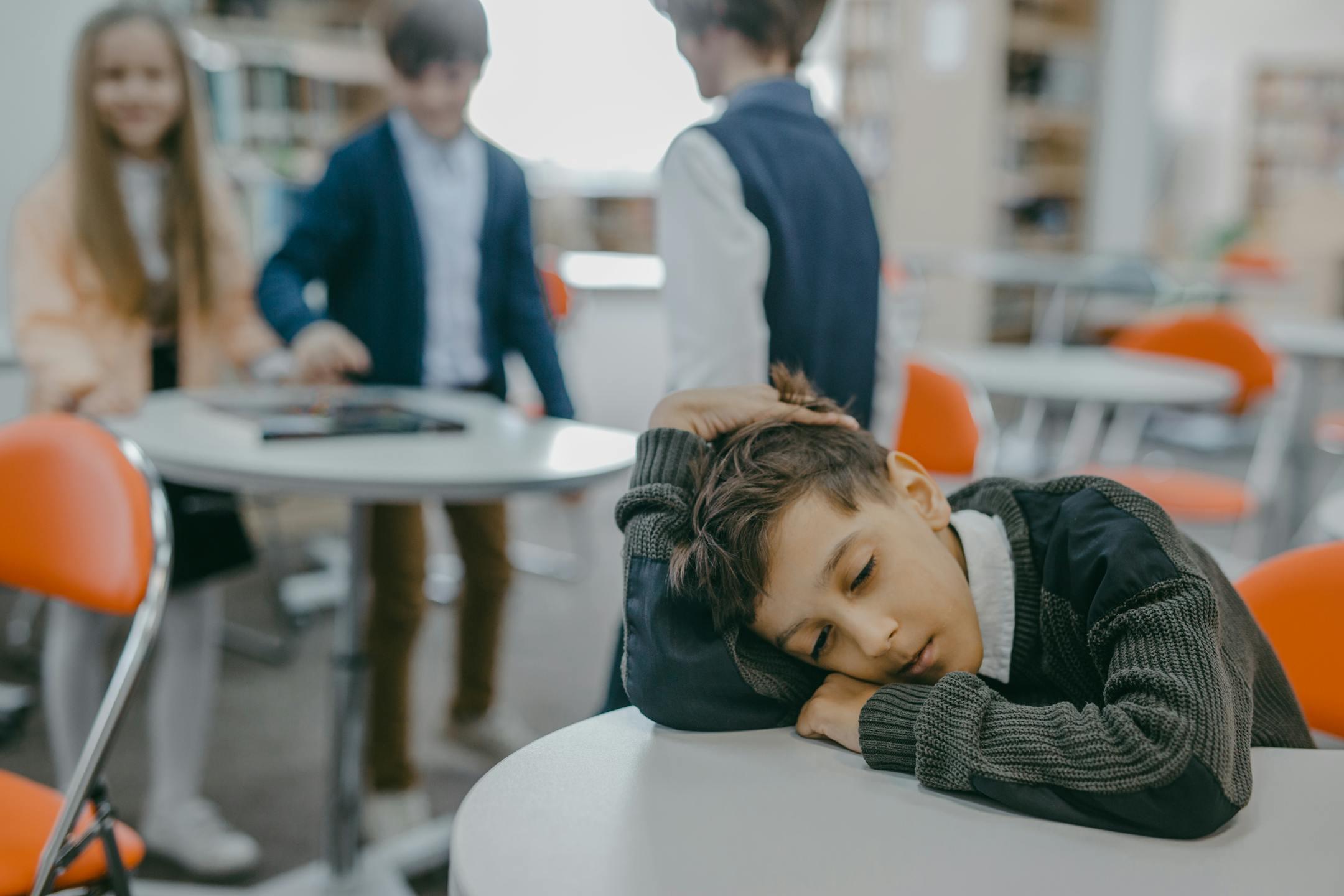 A sad child rests on a classroom desk while peers talk, symbolizing loneliness and school exclusion.