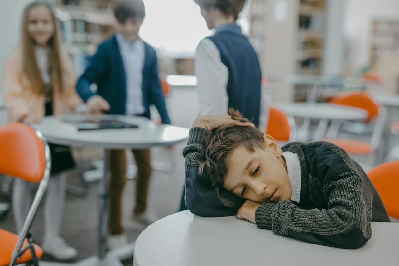A sad child rests on a classroom desk while peers talk, symbolizing loneliness and school exclusion.