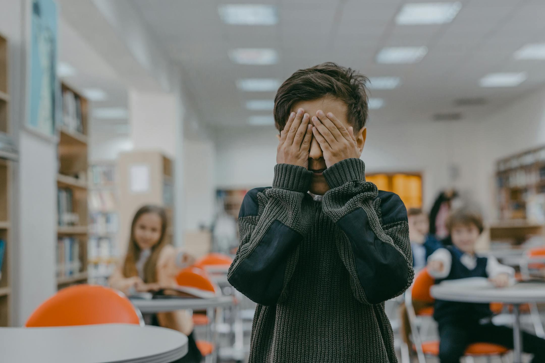 Young boy covers face in a bustling school classroom, indicating stress or anxiety.