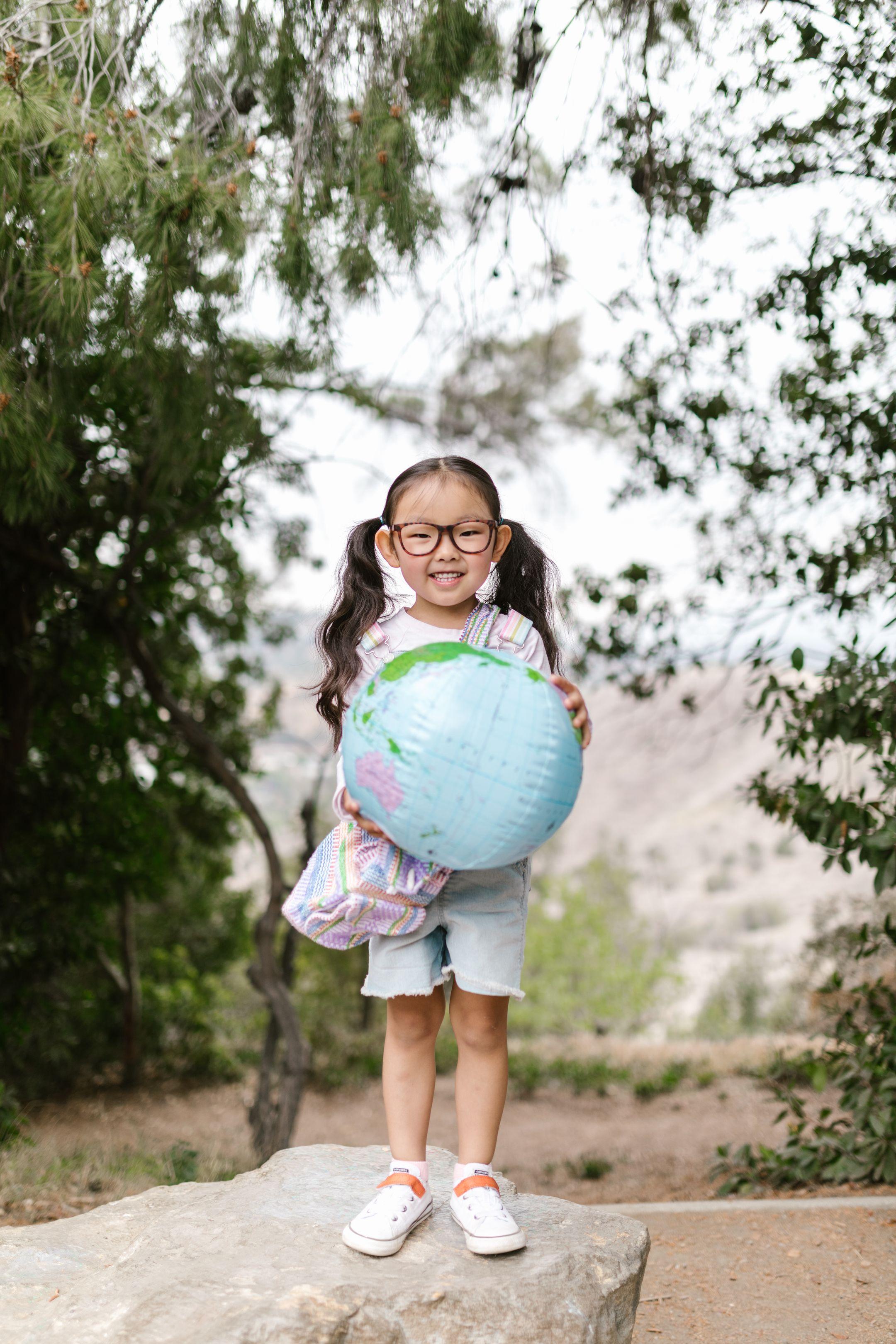 Smiling young girl with glasses holding a globe outdoors, symbolizing education and exploration.