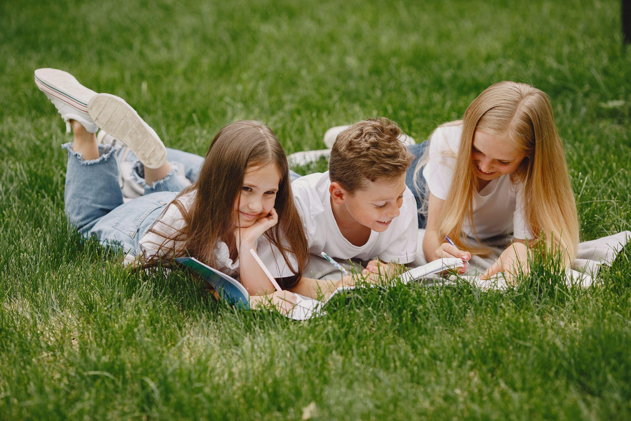 Three children lying on grass reading and smiling outdoors.
