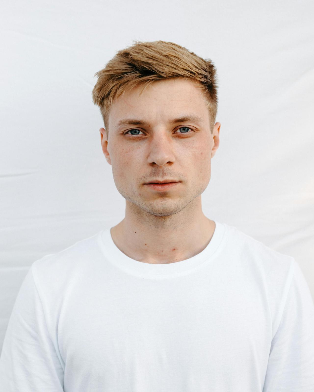 Close-up portrait of a young man with blue eyes and a neutral expression wearing a white t-shirt.