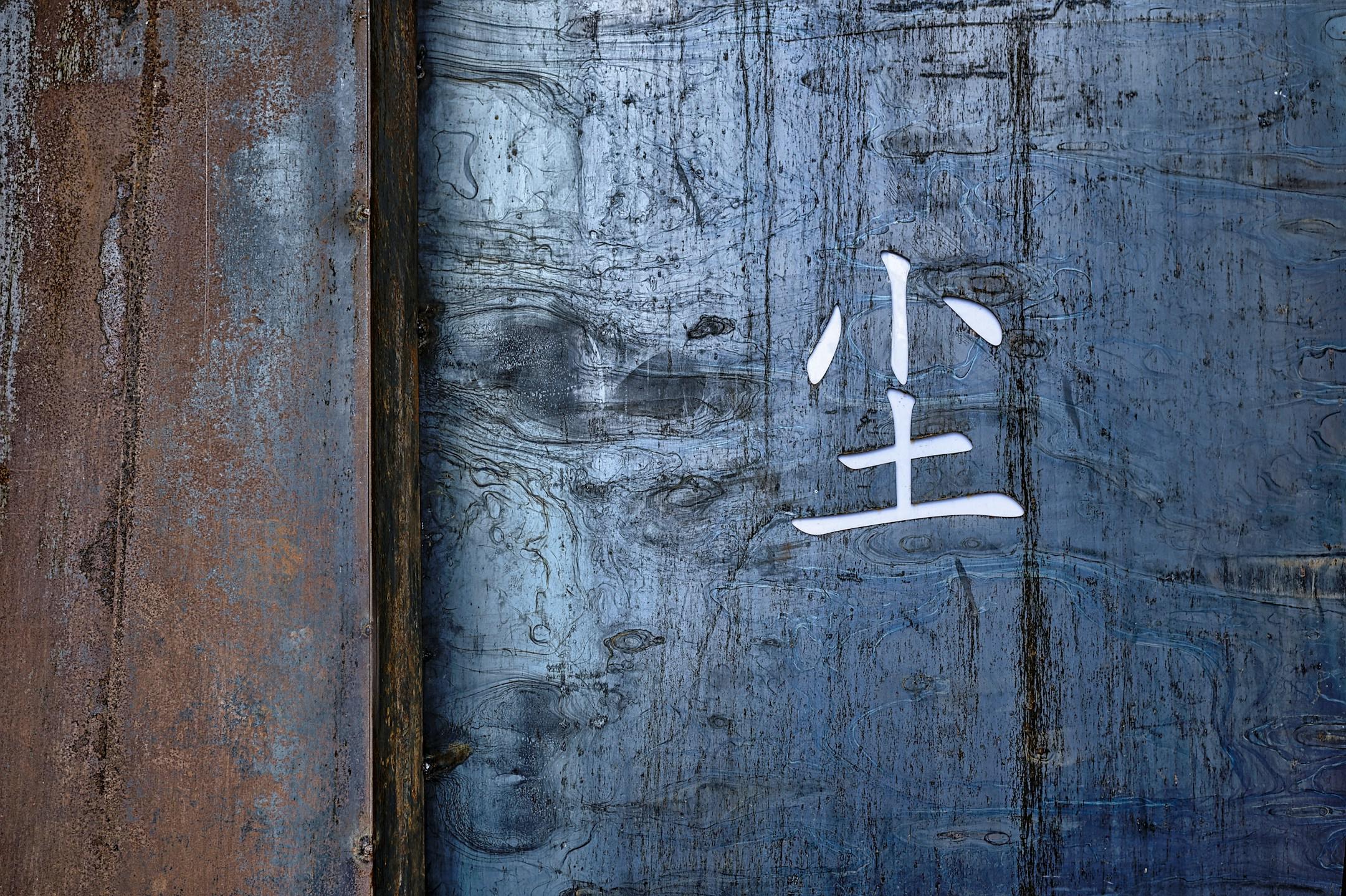 Rusted metal Wall With Chinese Symbol meaning "DUST" Painted In Asian Characters - A rustic metal wall exhibits a blend of textures and colors, showing deep blue and brown tones. A white symbol, likely of Asian origin, is prominently painted on the surface, adding an artistic touch to the weathered metal. The overall ambiance reflects a sense of age and cultural significance.