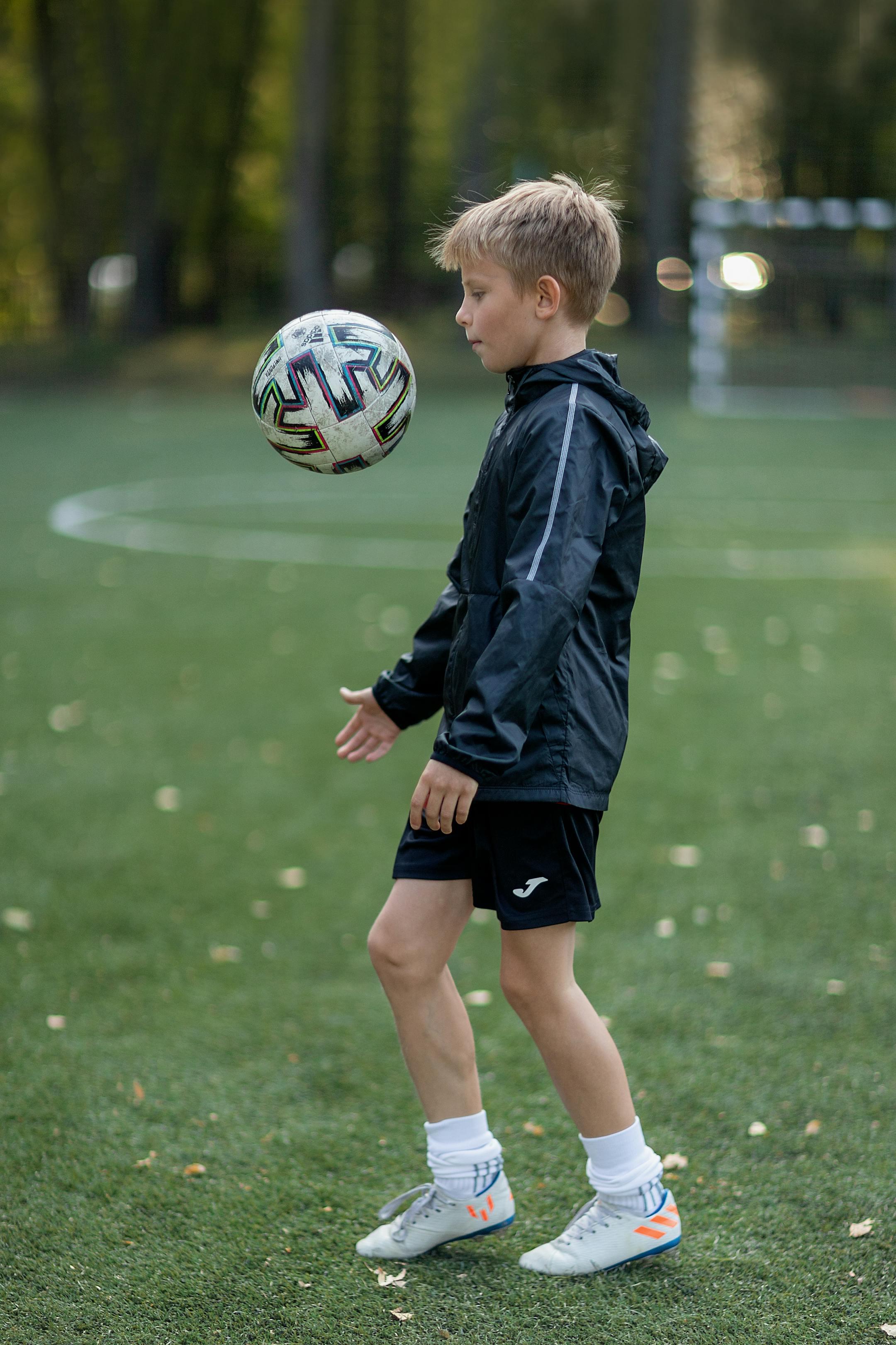 A young boy in sportswear practices soccer skills on a green football field during the day.