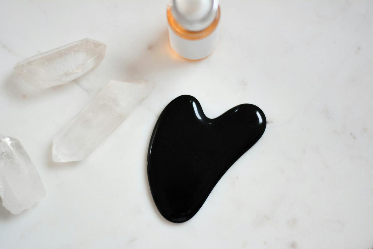 Black gua sha massage tool on a marble surface surrounded by crystals and facial serum.