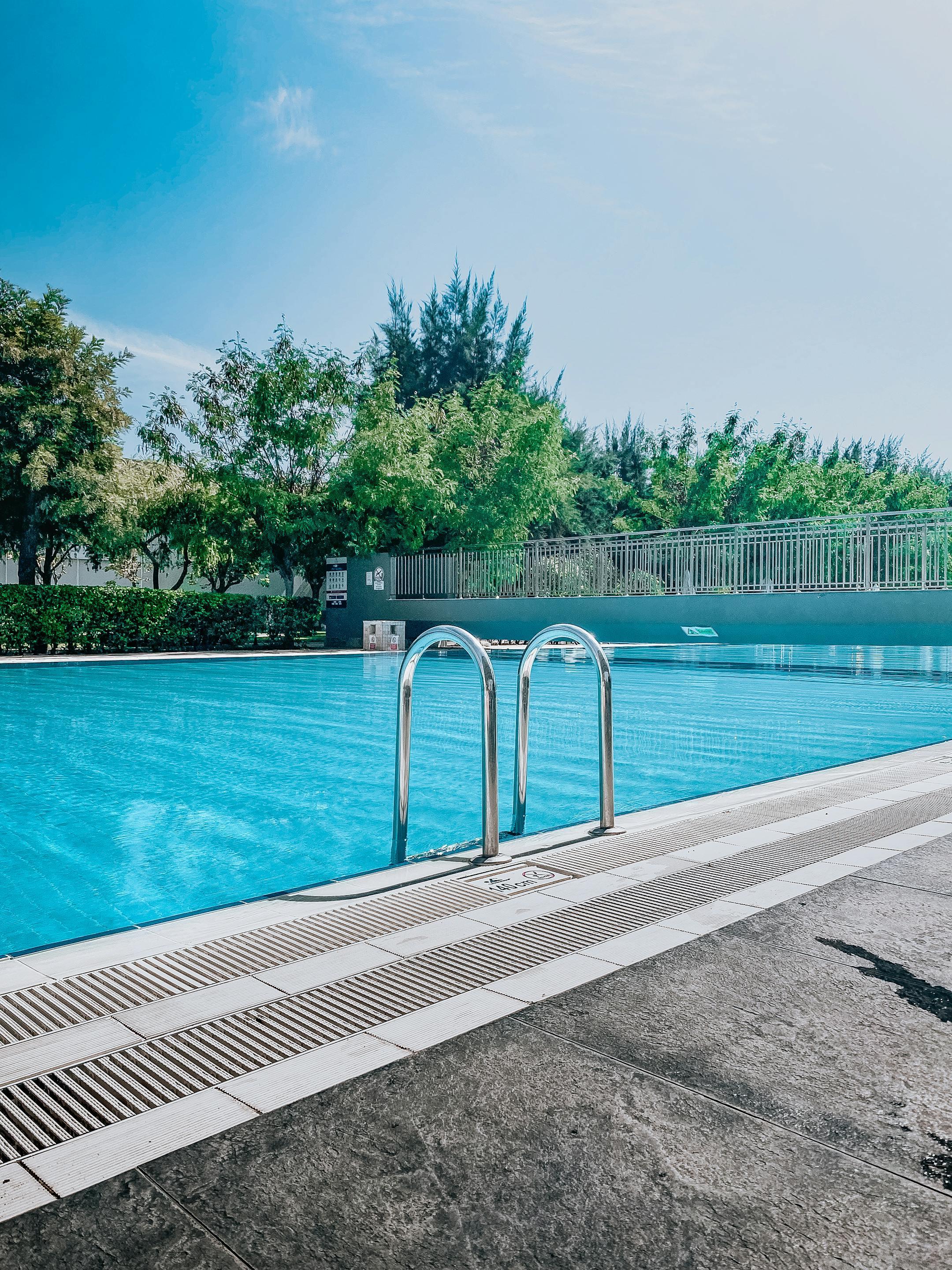 A serene outdoor swimming pool with clear blue water and lush greenery under a bright sky.