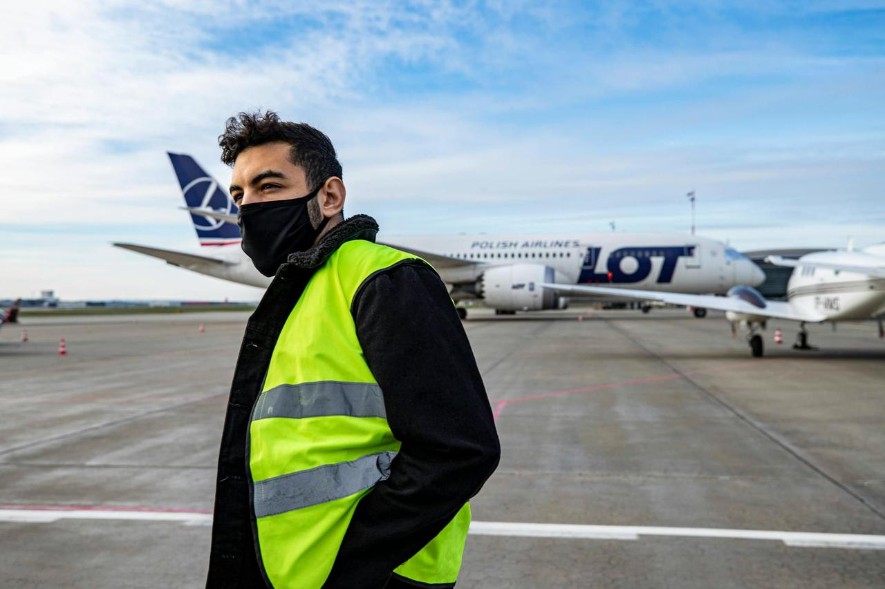 Man at airport runway wearing a reflective vest and face mask near LOT Polish Airlines plane.
