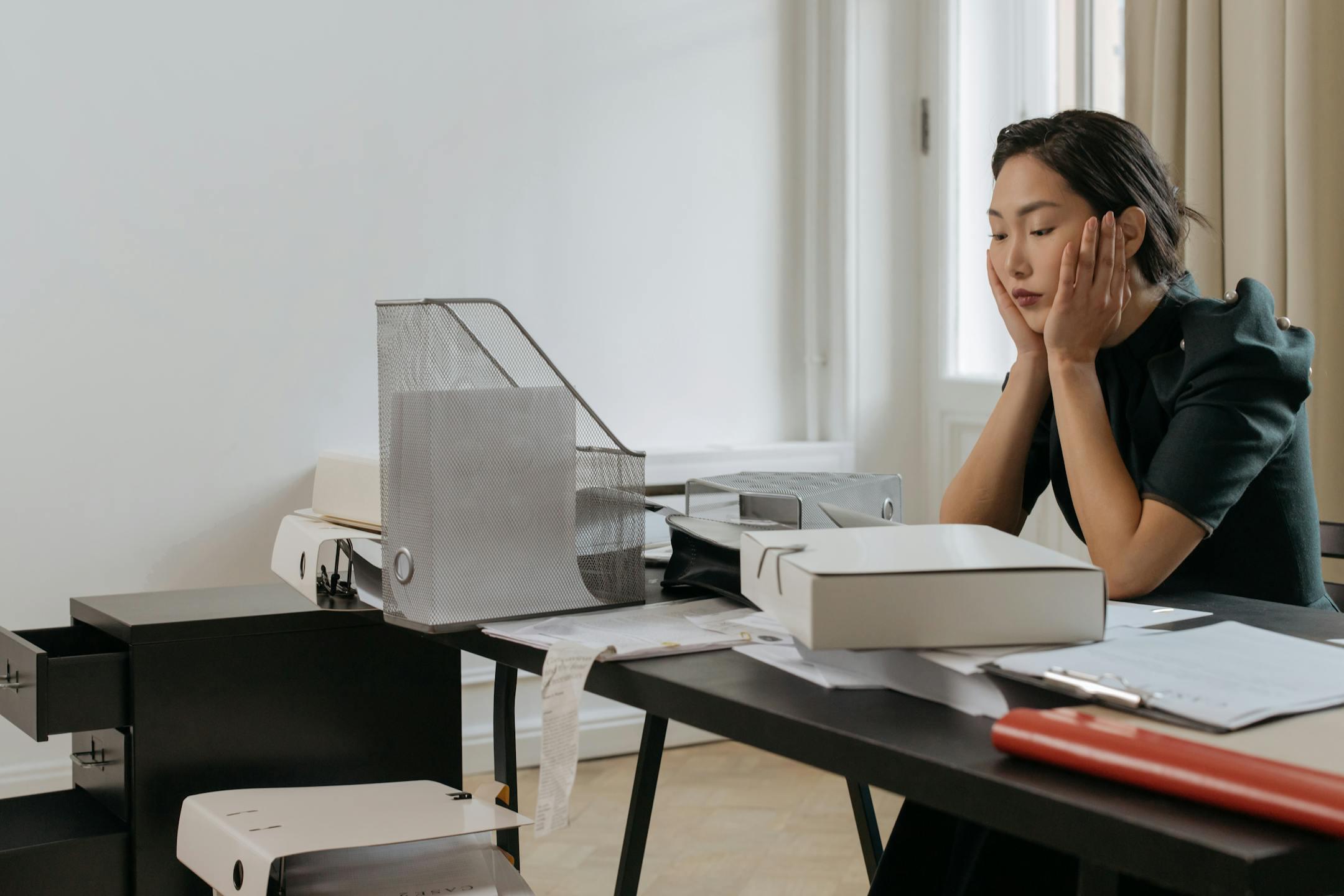 Young woman looks stressed while surrounded by documents at her office desk.
