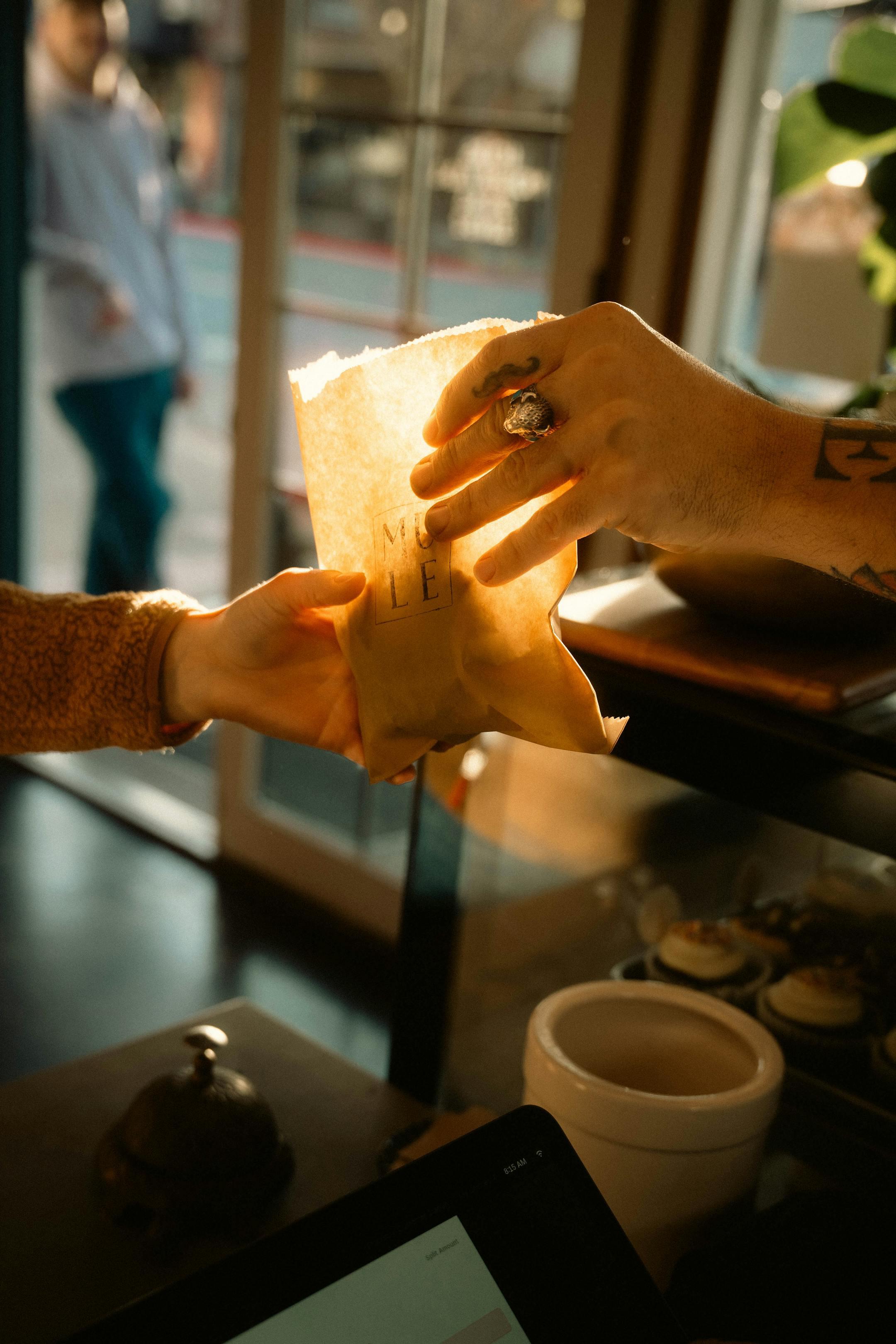 A person hands over a brown paper bag to another in a cozy café setting with warm lighting.
