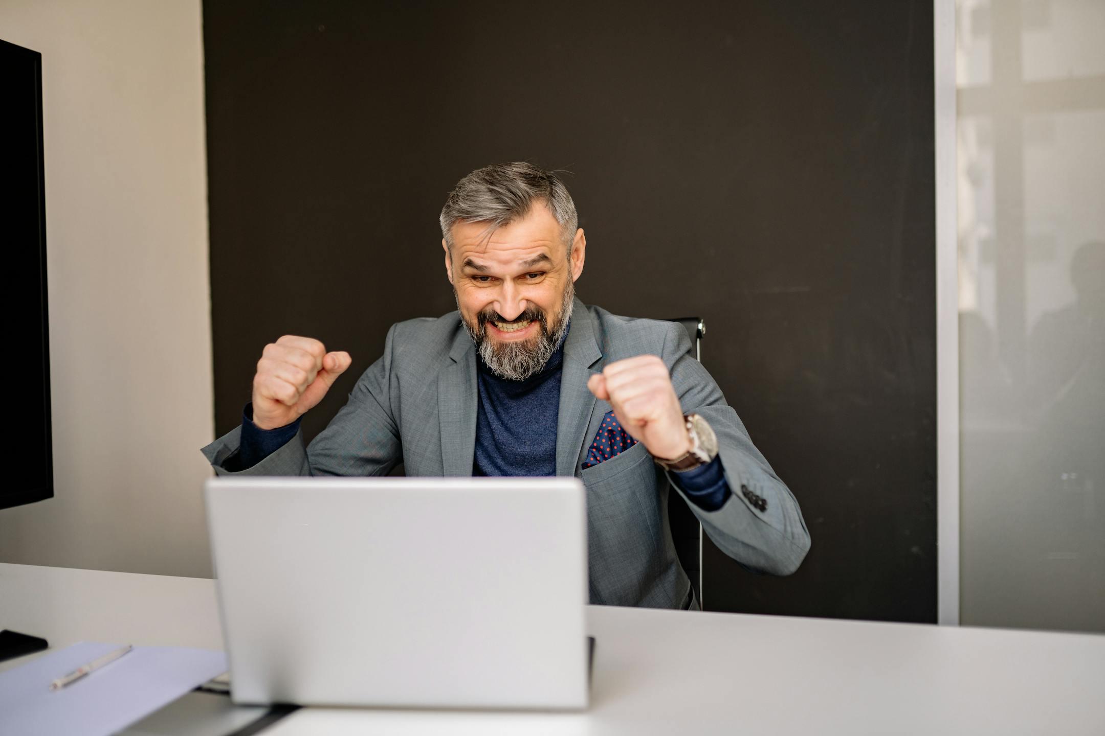 Happy businessman celebrating success while looking at laptop in modern office.