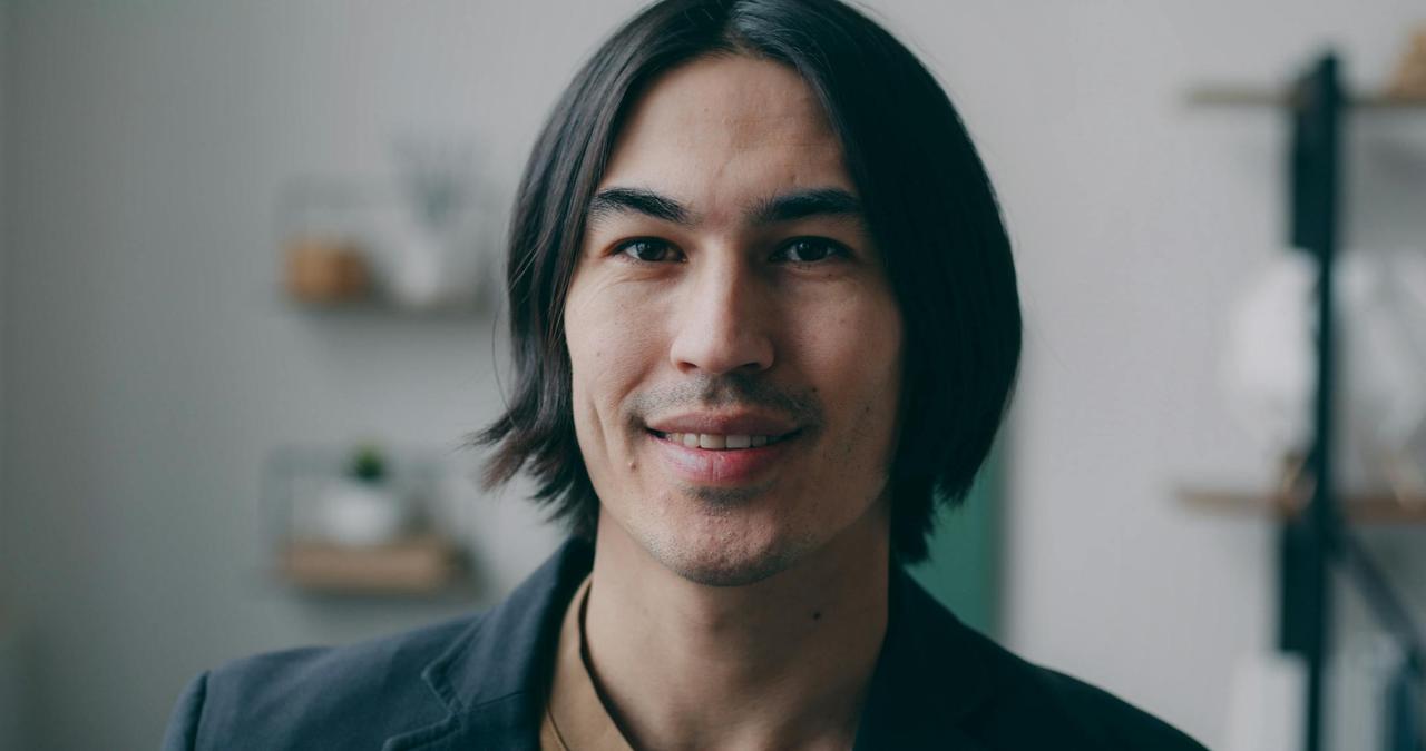 Close-up portrait of a smiling man with long hair indoors.