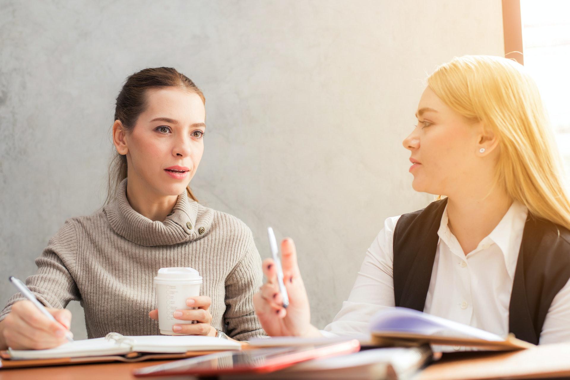 Two women providing real estate investing coaching at a table, one of them holding a cup of coffee and speaking with the other one listening.