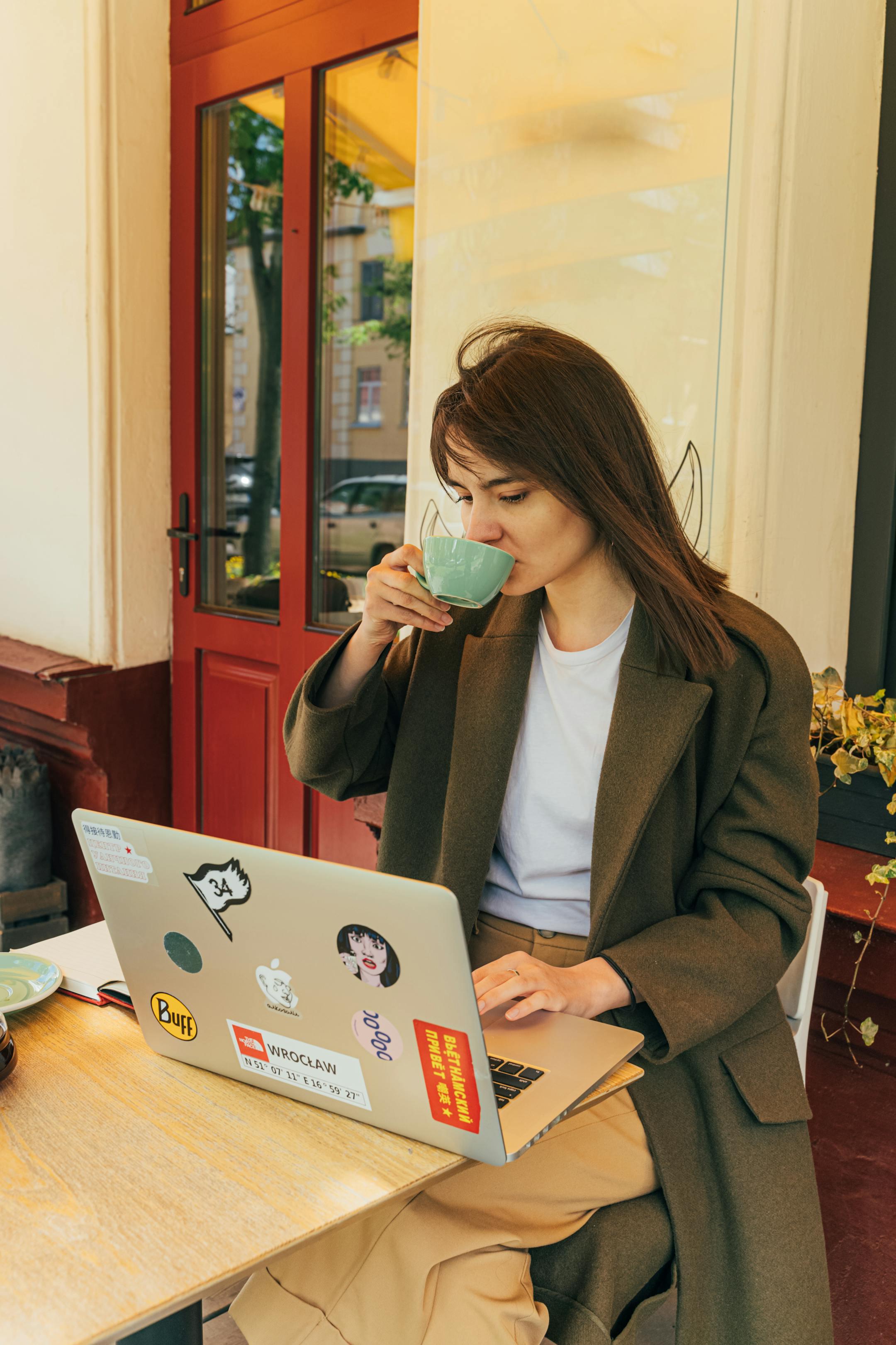 Woman in a coat drinking coffee and working on a laptop at an outdoor café.