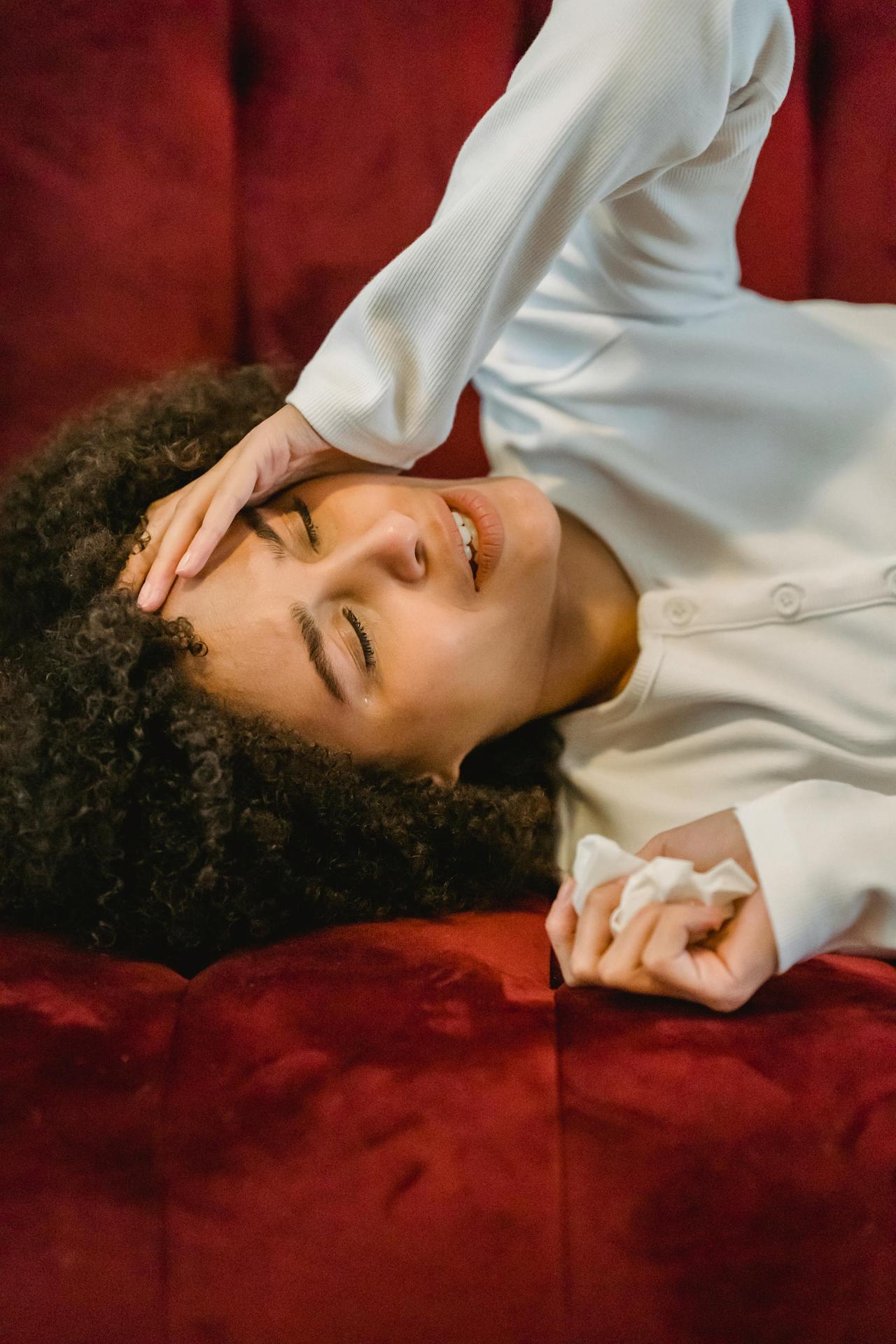 Unhappy African American female with closed eyes touching forehead while lying on comfortable sofa with tissue in cozy room at home