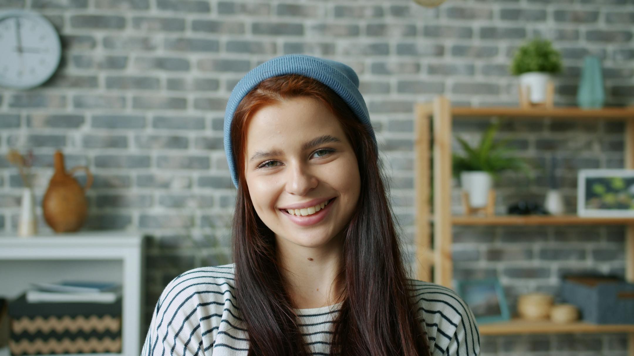 A happy young woman wearing a beanie smiles in a cozy indoor setting with a brick wall backdrop.