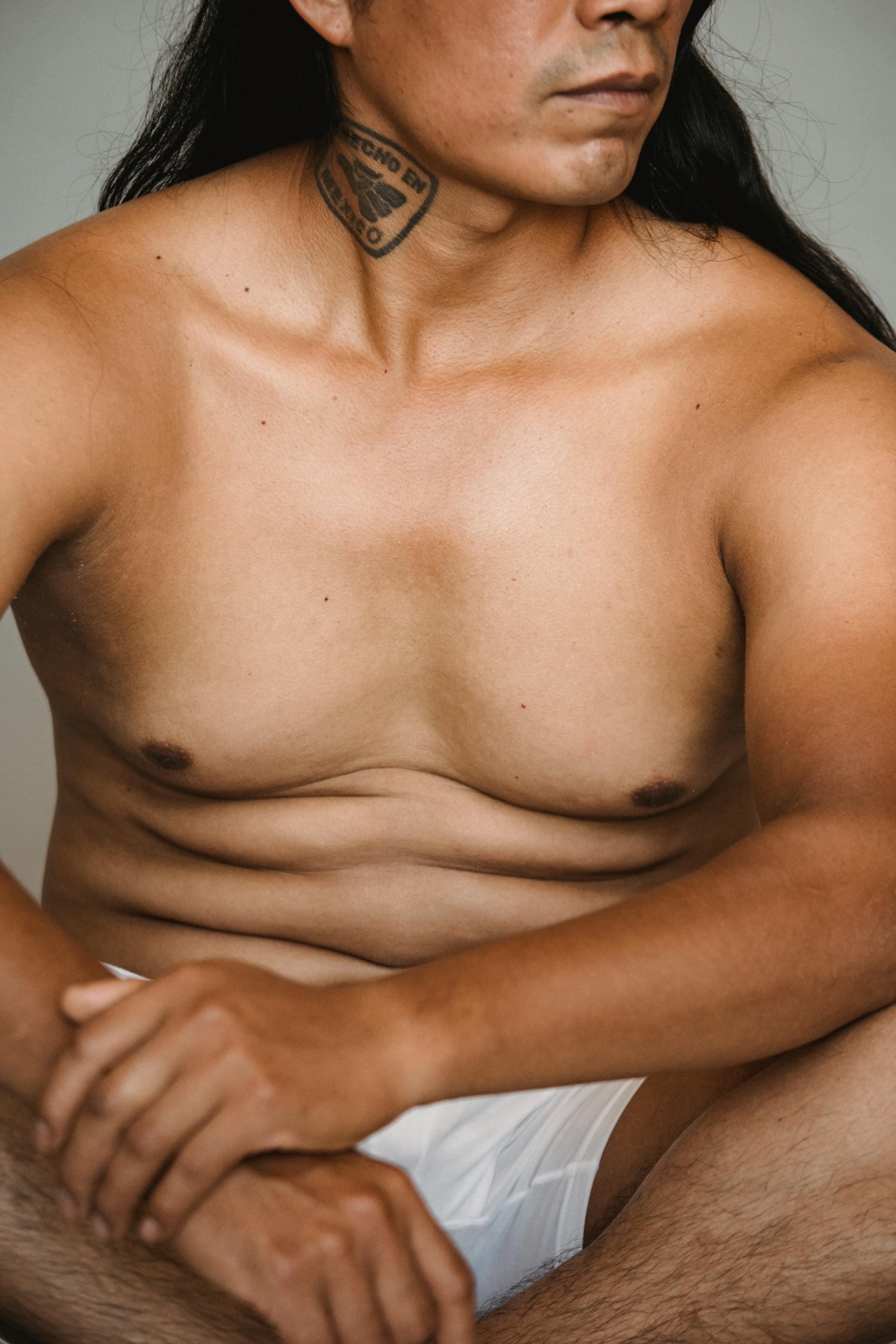 Portrait of a shirtless tattooed man sitting indoors, showcasing masculinity and body art.