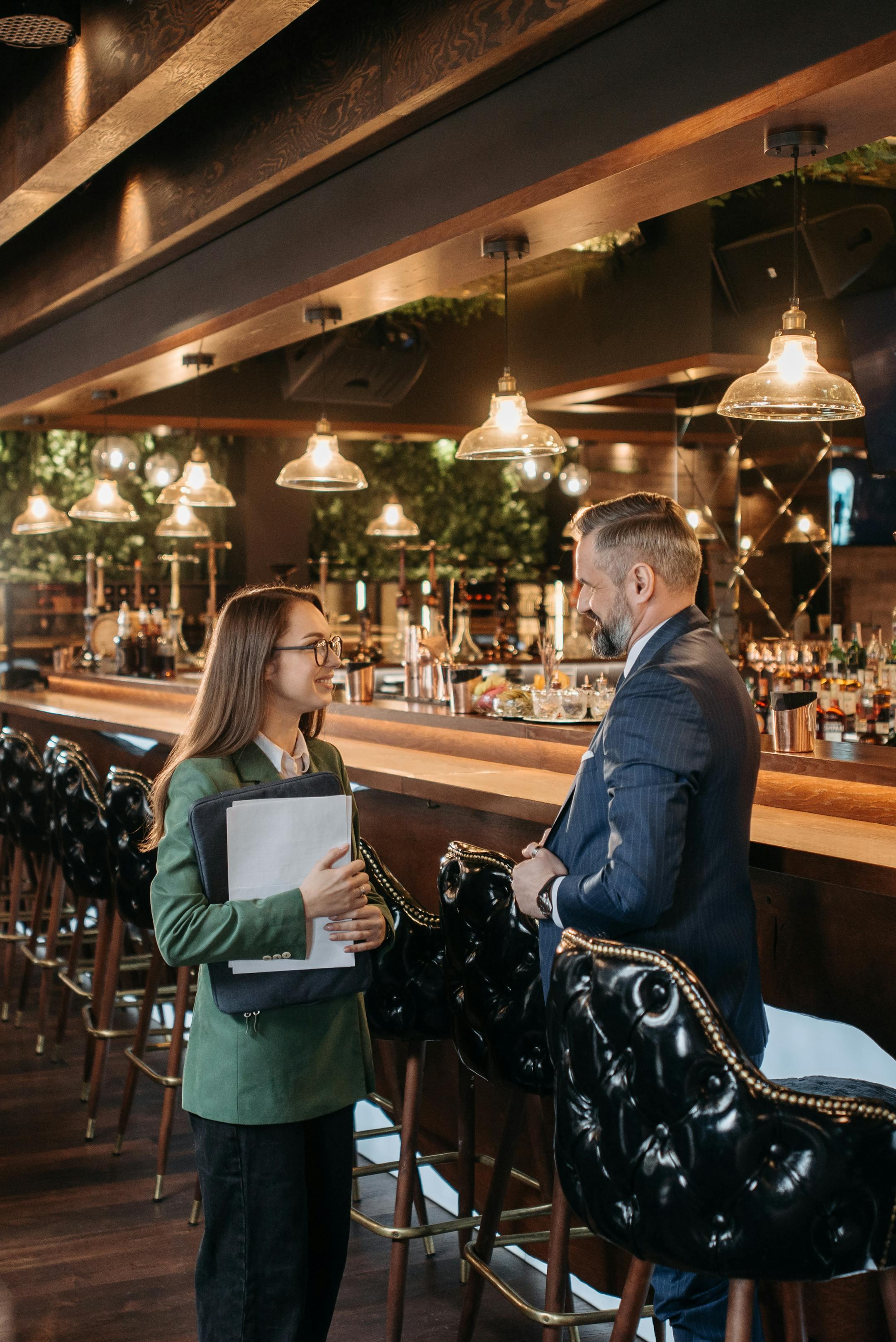 Two professionals engage in a business discussion at a stylish bar with modern design elements.