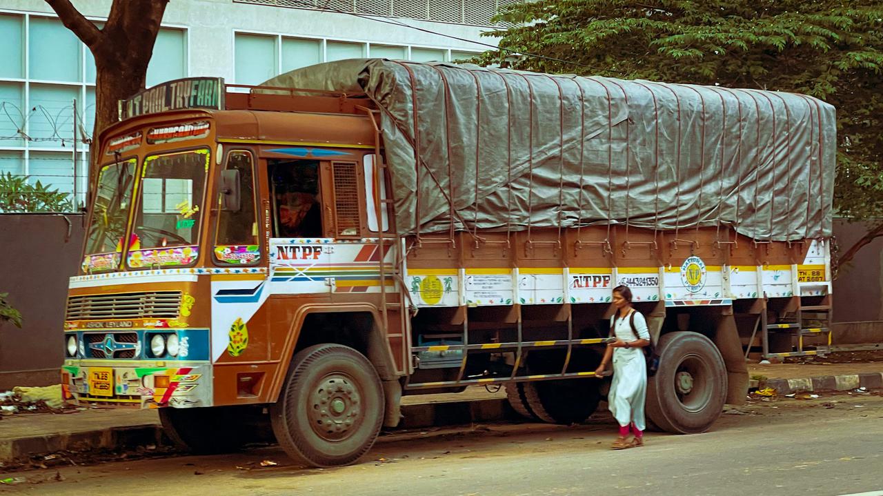 A colorful cargo truck decorated with traditional Indian motifs is parked on a city street. The truck, covered with a tarp, is used for transporting goods. In the foreground, a woman walks by, and a cyclist passes on the road. The scene captures everyday urban life in India, showcasing the coexistence of pedestrians, vehicles, and local businesses. The background includes a building with barbed wire and trees, adding to the urban environment's dynamic and bustling atmosphere.