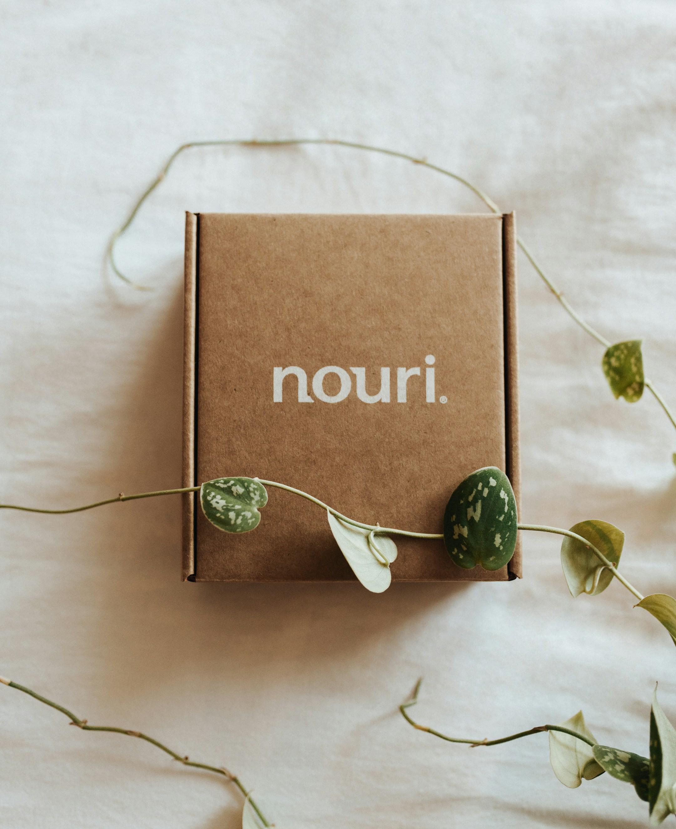 Package of Nouri, a proprietary probiotic and omega blend, on white comforter surrounded by beautiful vines of green leaves.
