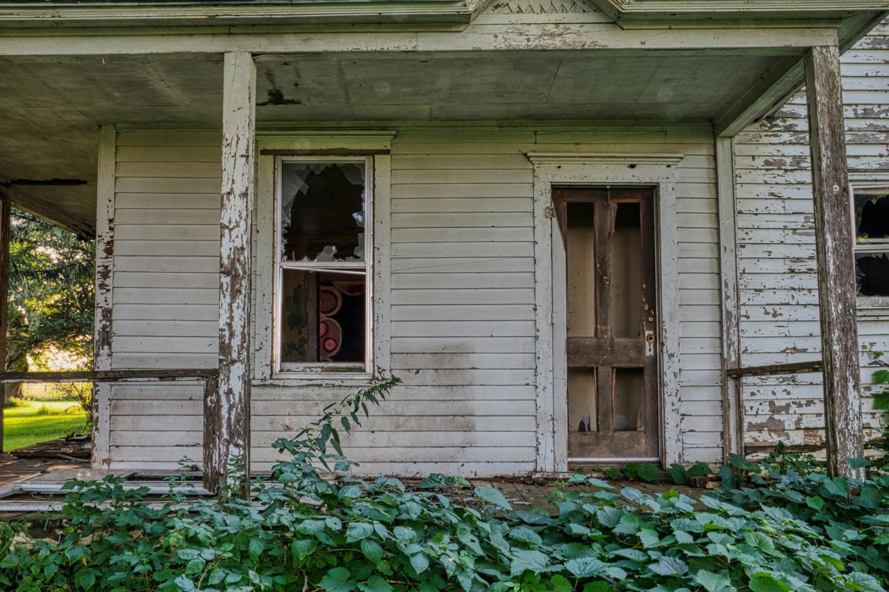 An old abandoned house with broken windows, wooden door, surrounded by greenery, showcasing vintage architecture.