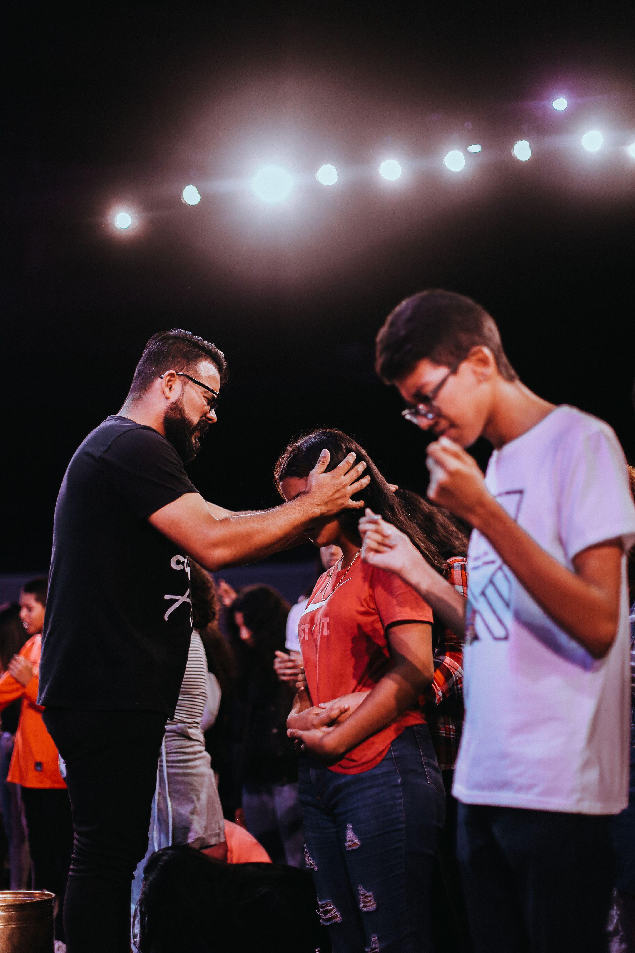 A group of young people gathered indoors for a prayer session under warm lights.