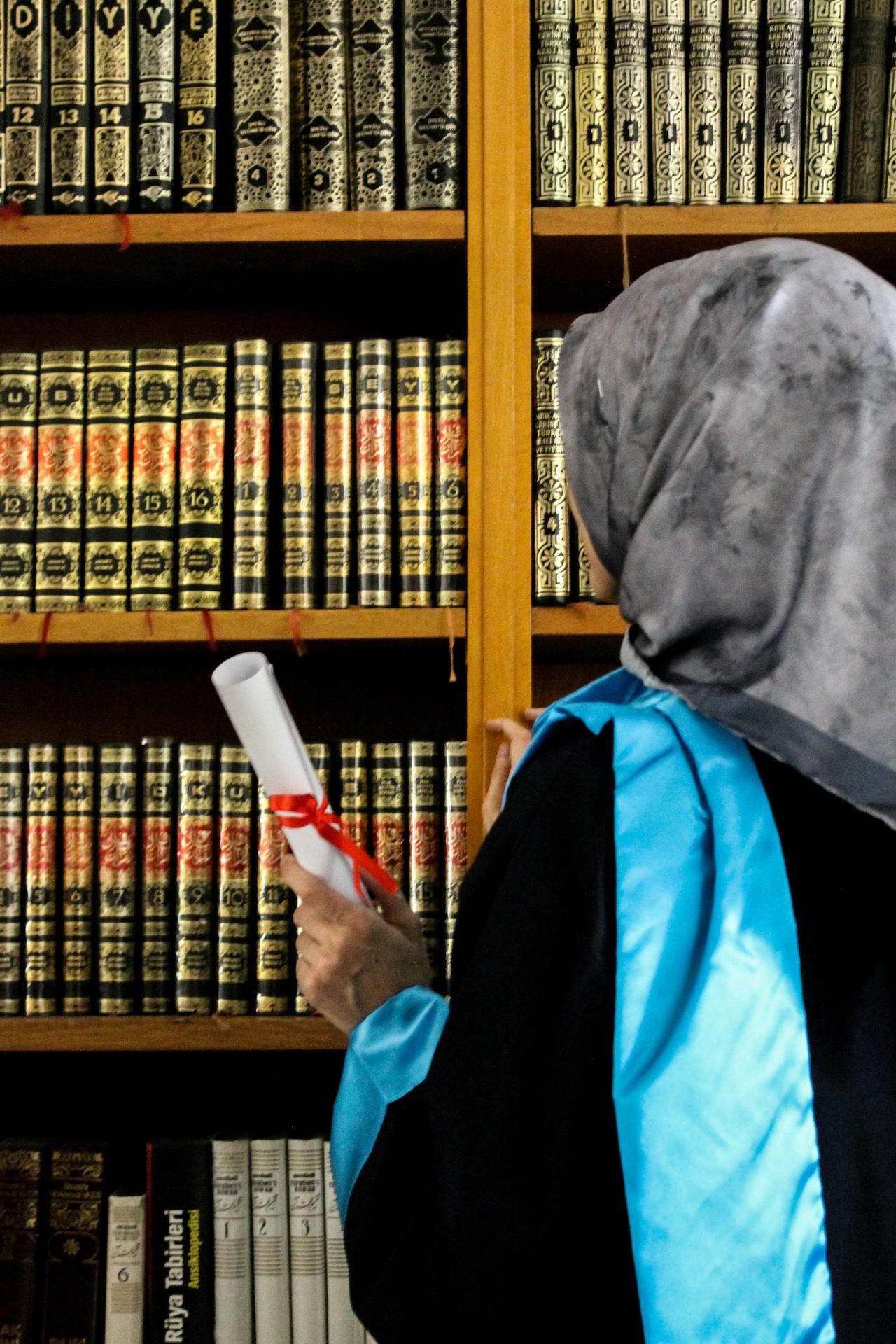 A woman in a hijab holding a diploma, standing in a library beside a bookshelf full of books.