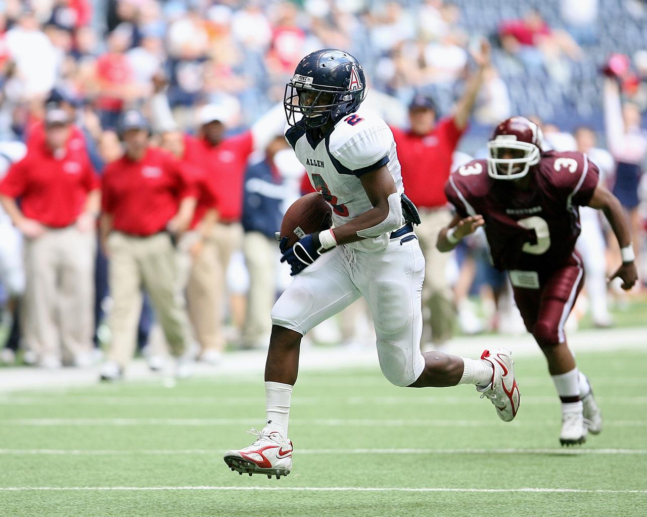 High school running back on his way to scoring a touchdown with coaches and fans looking on.