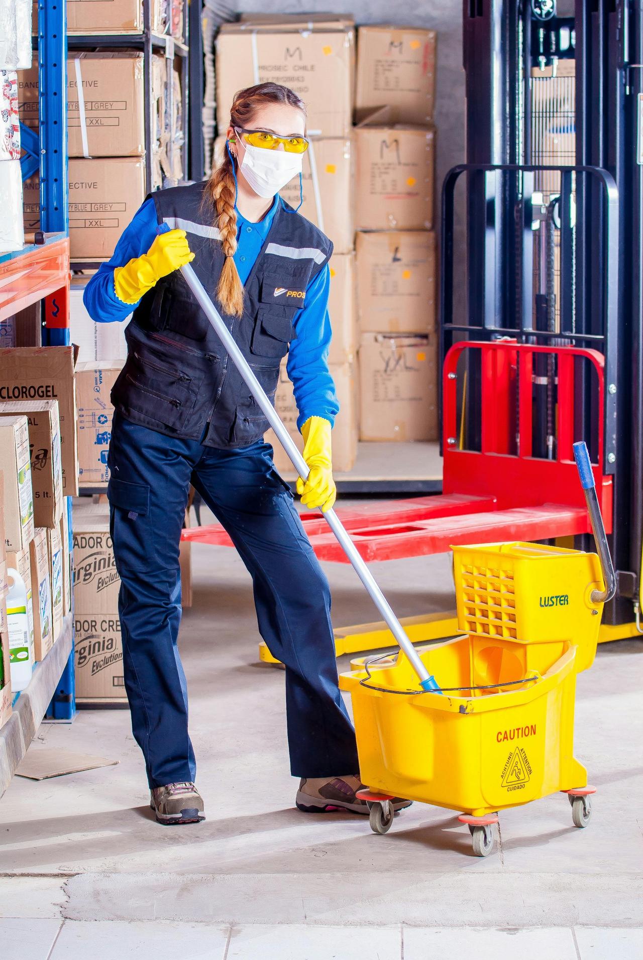 Female janitor in safety gear cleaning a warehouse floor with a mop and bucket.
