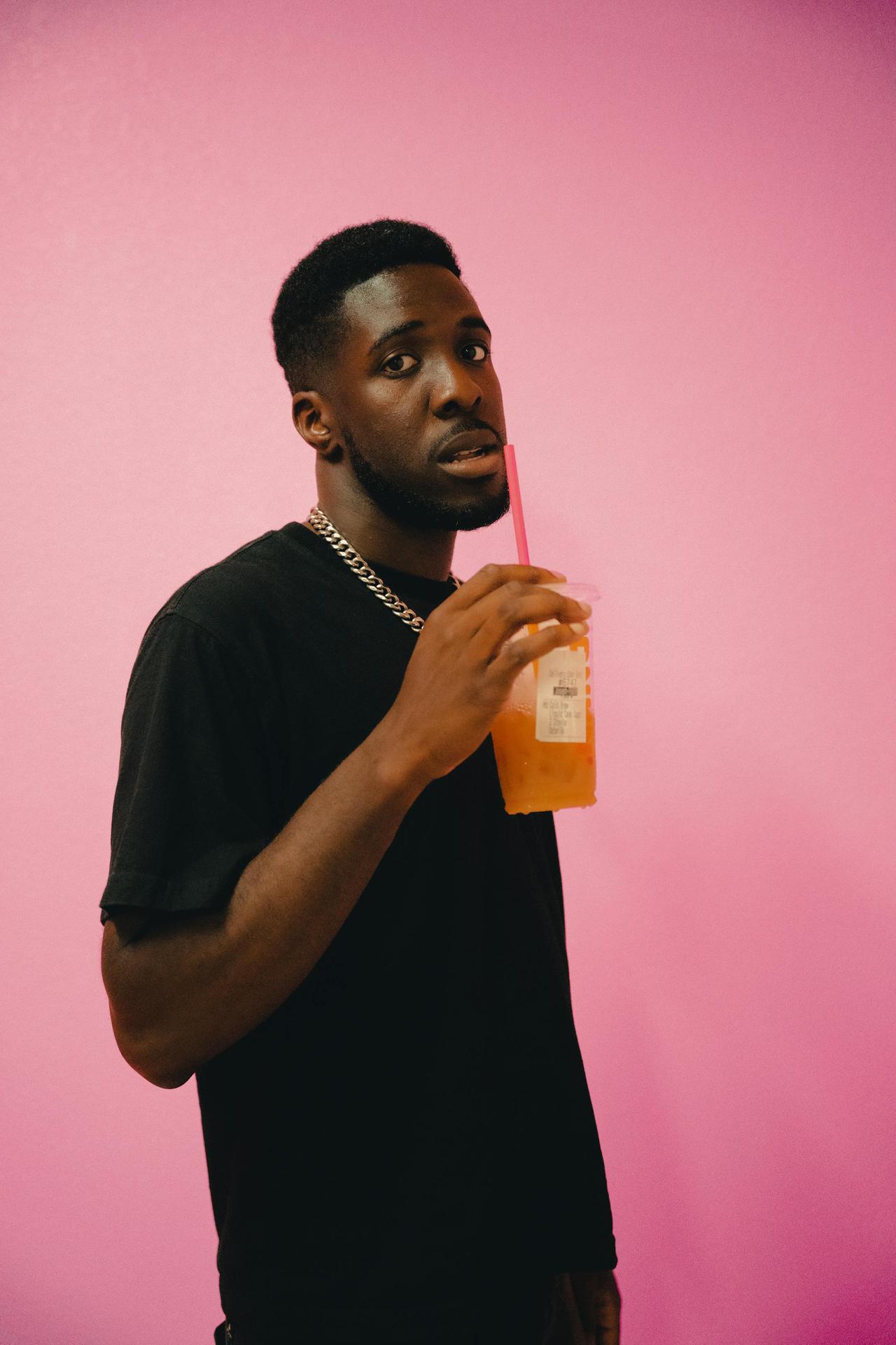 Adult man sipping juice through a straw in a studio with pink background, conveying a casual and refreshing mood.