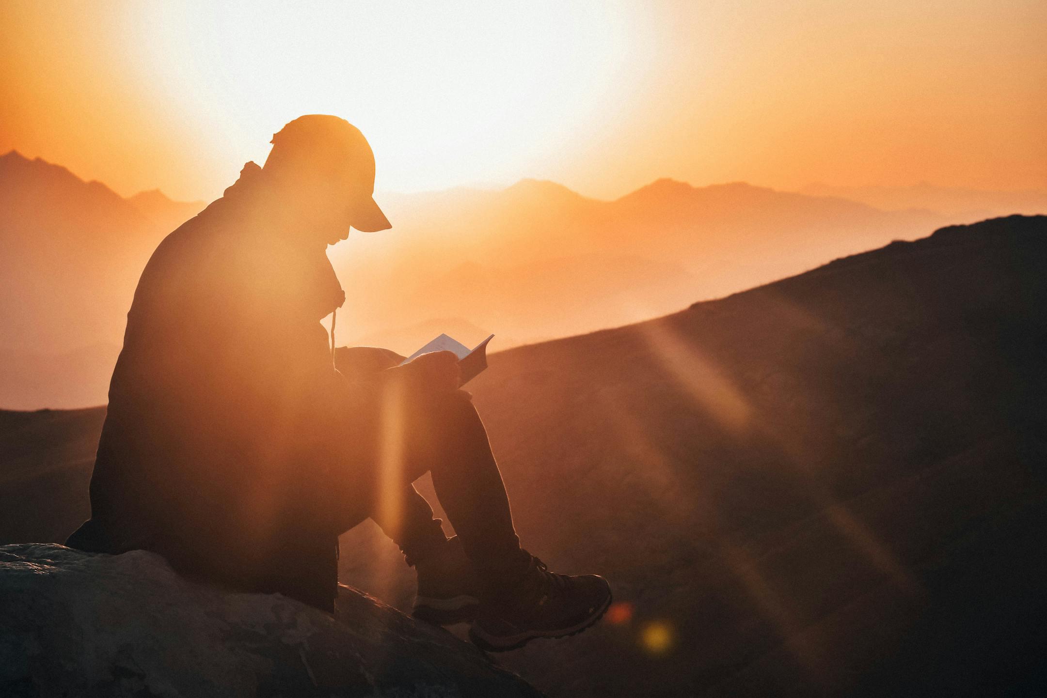 A man reads a book silhouetted against a vibrant sunset, seated on a mountain edge.