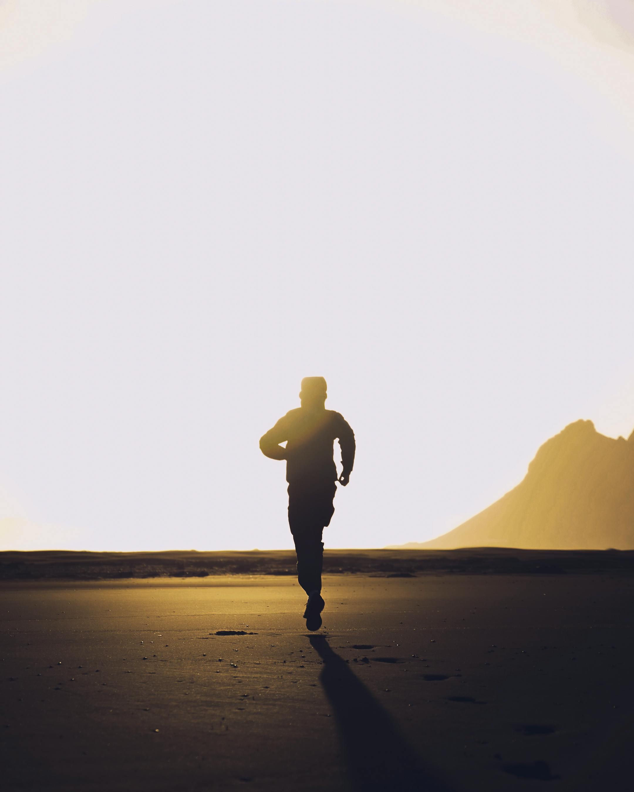 Back view of silhouette of anonymous athletic male running on dark sandy beach at sunset
