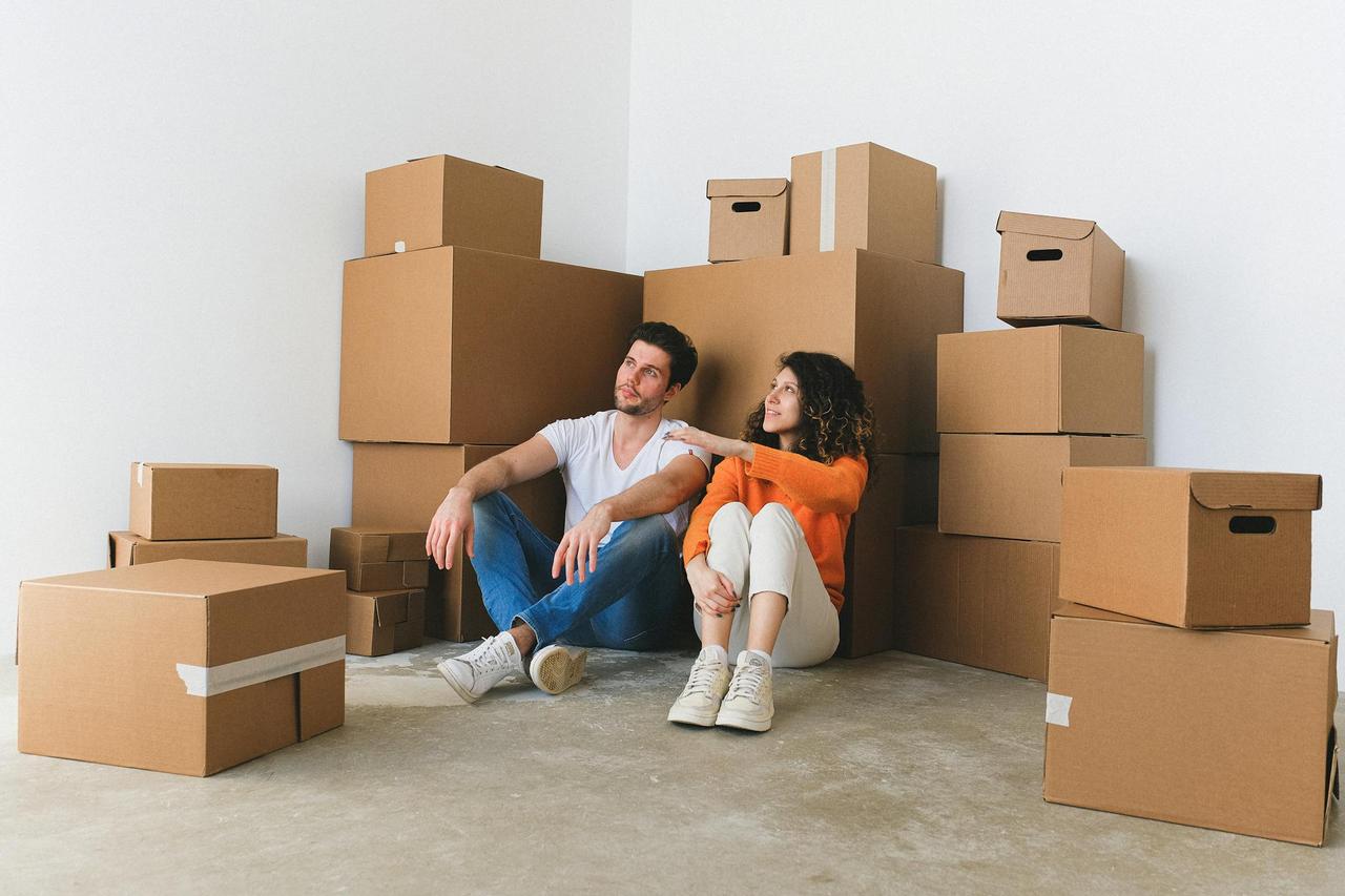 Full body of young happy couple in casual clothes sitting on floor leaning on stack of cardboard boxes and chatting after relocation in new flat