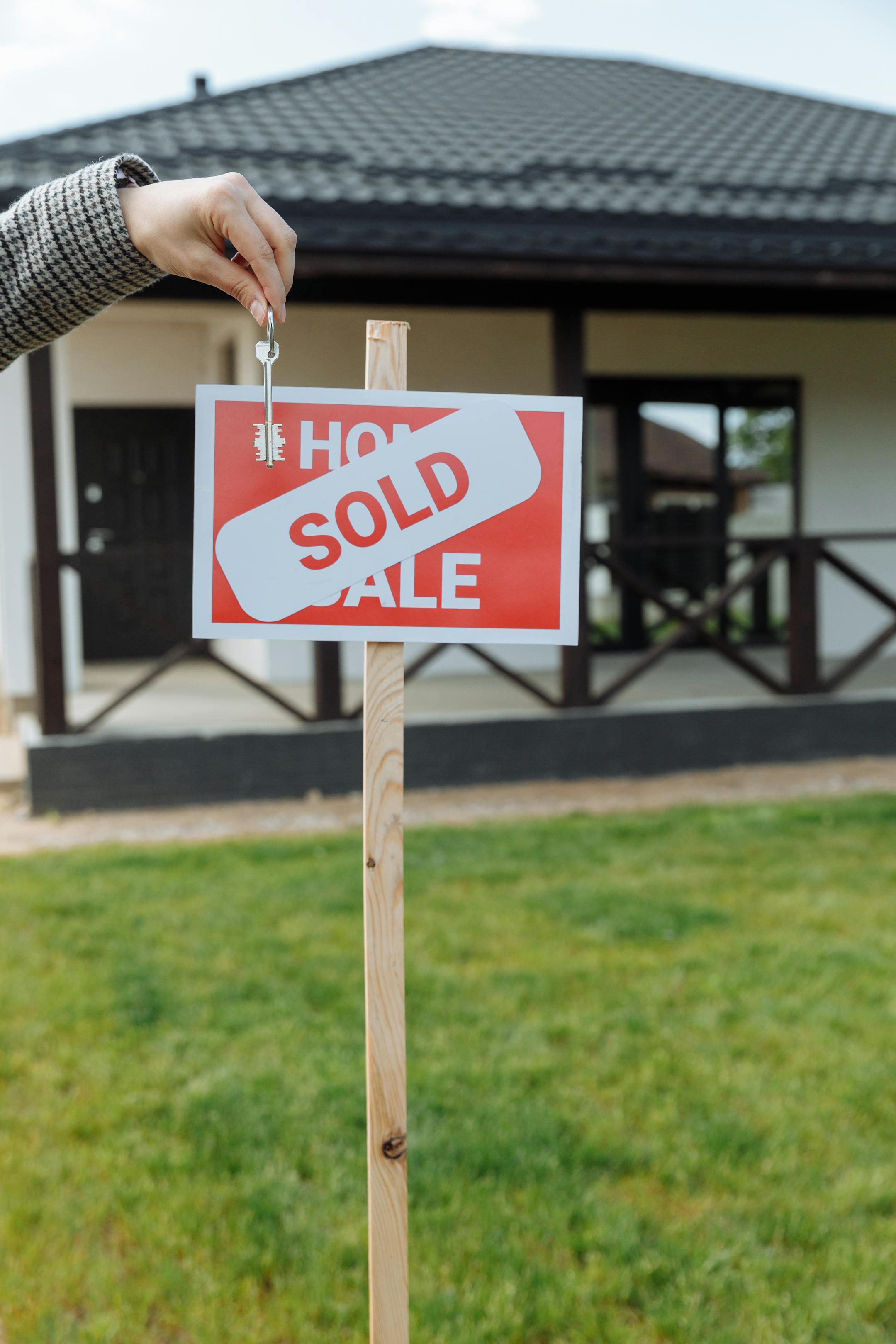 A hand holding a key in front of a sold sign and a house, symbolizing a successful real estate transaction.