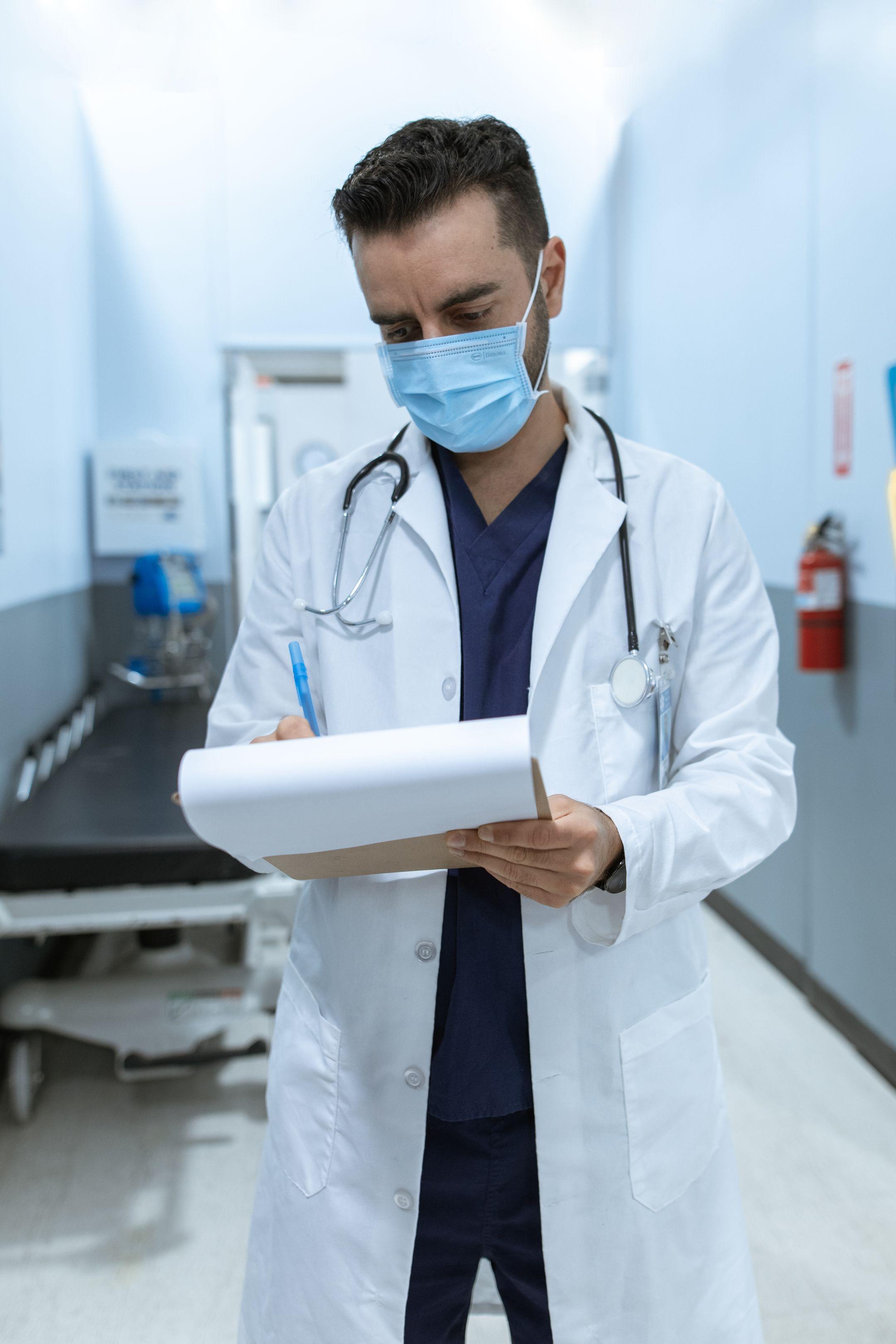 Medical professional wearing mask, writing on chart in hospital hallway.