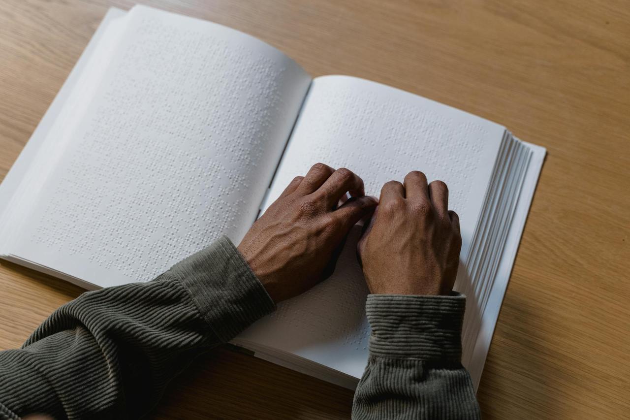Hands reading a braille book on a wooden table, showcasing assistive technology for the visually impaired.