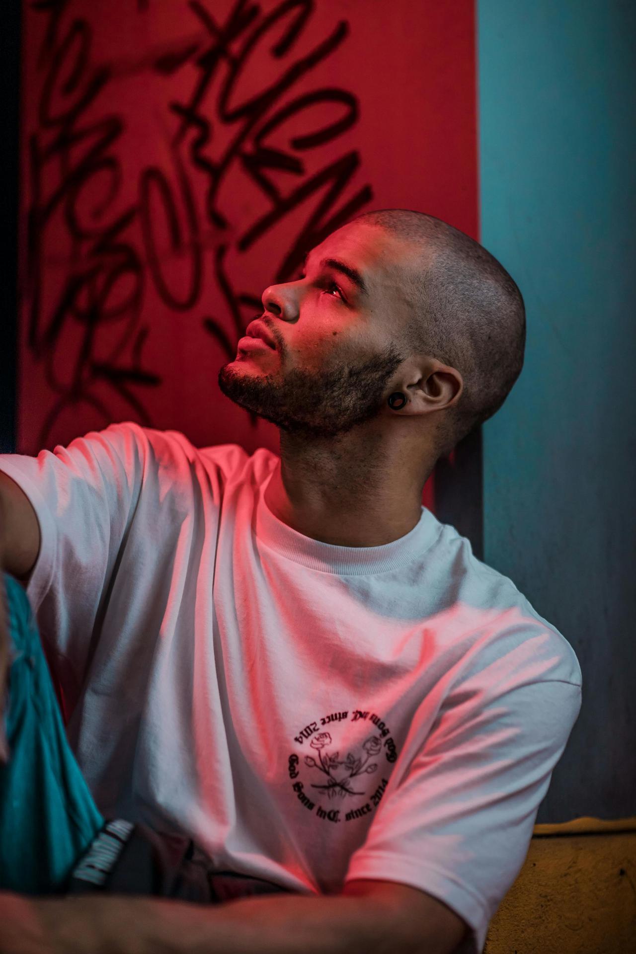 Portrait of a bald man with facial hair sitting indoors against graffiti, bathed in red and blue neon light.