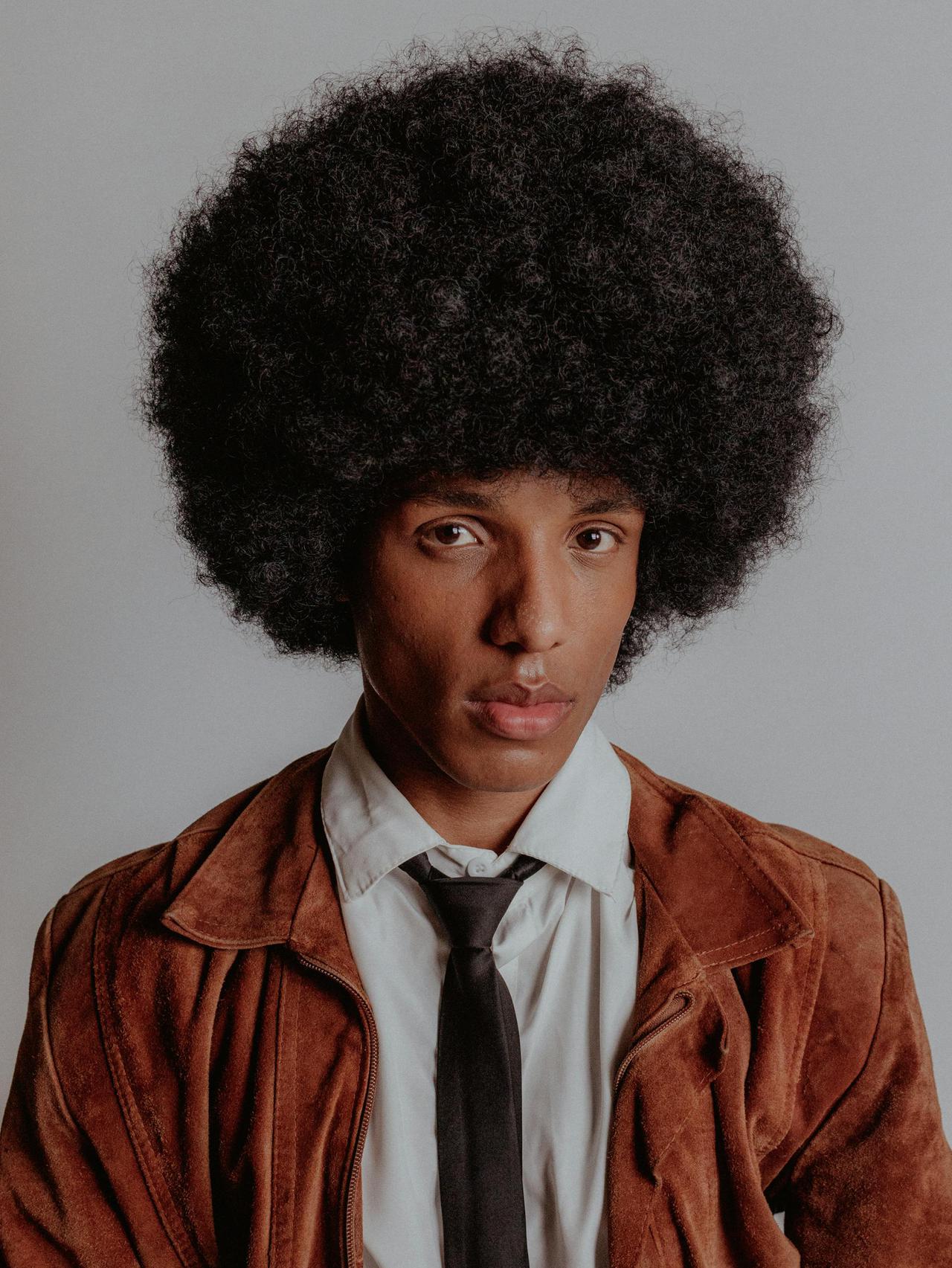 Young man in brown jacket with an afro hairstyle poses against a neutral background.