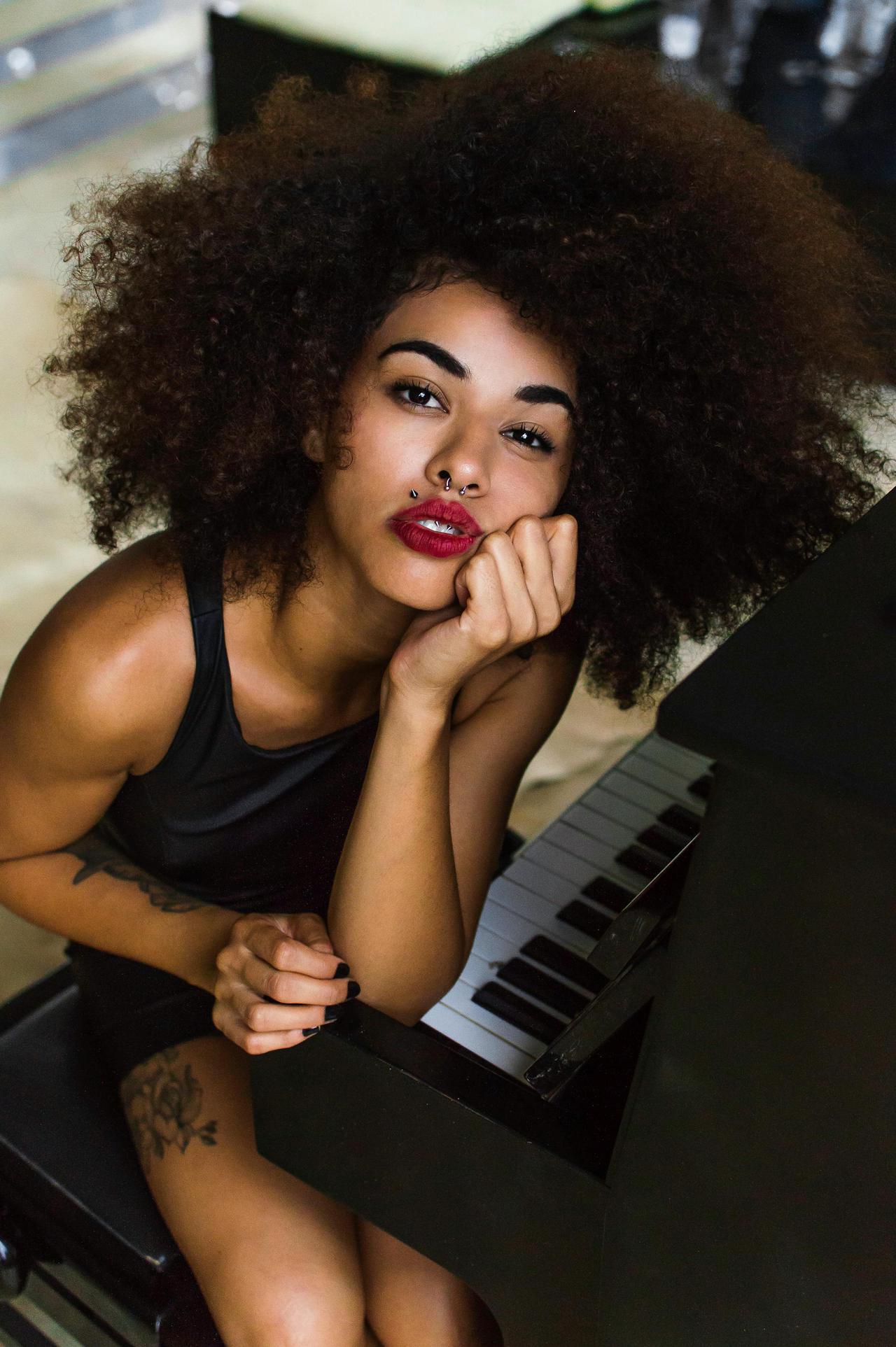 A beautiful woman with curly hair poses at a piano, featuring tattoo and bold red lips.
