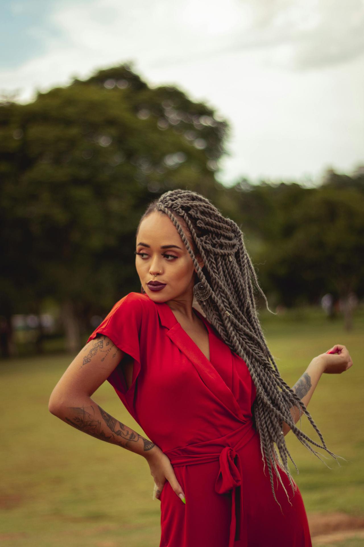 Confident woman with braided hair poses in a vibrant red dress in a park setting.