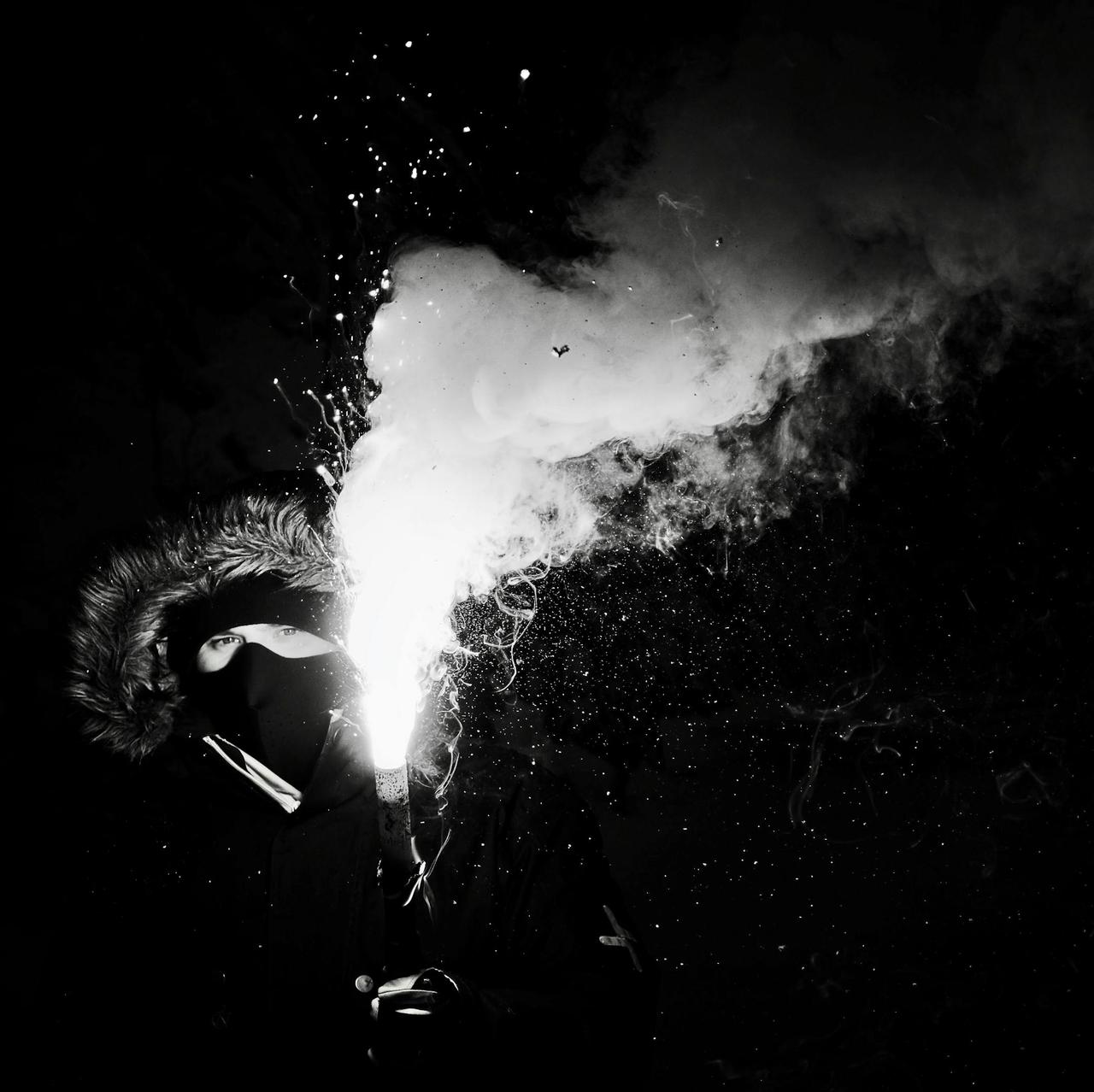 Black and white photo of a masked person holding a flare with smoke at night.