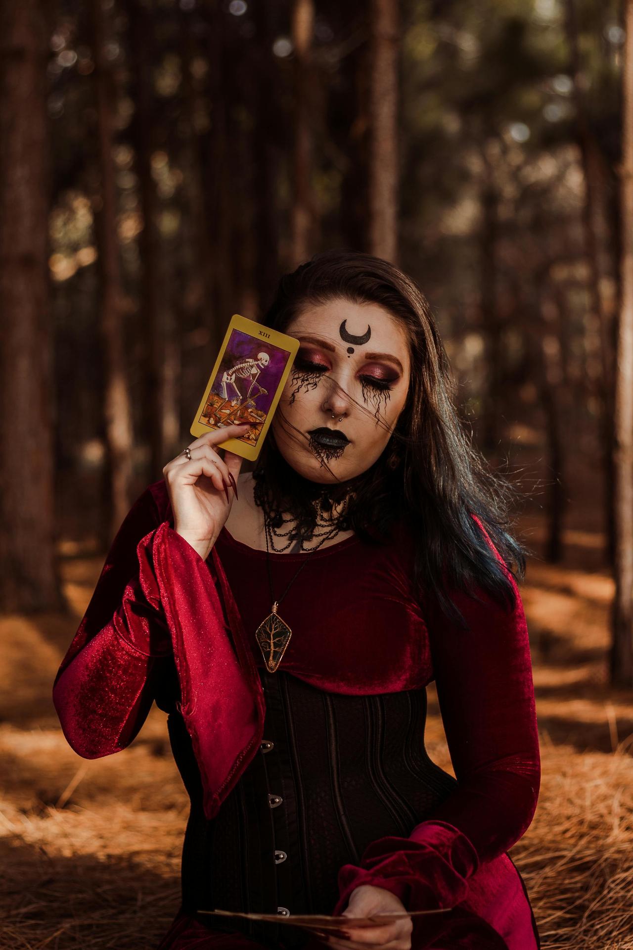 Enigmatic woman in gothic attire holds tarot card amidst autumn woods.