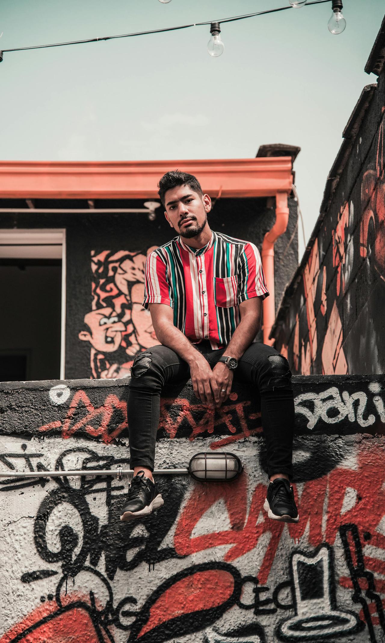 Young man with facial hair in colorful shirt sitting on graffiti wall in Medellín, Colombia.