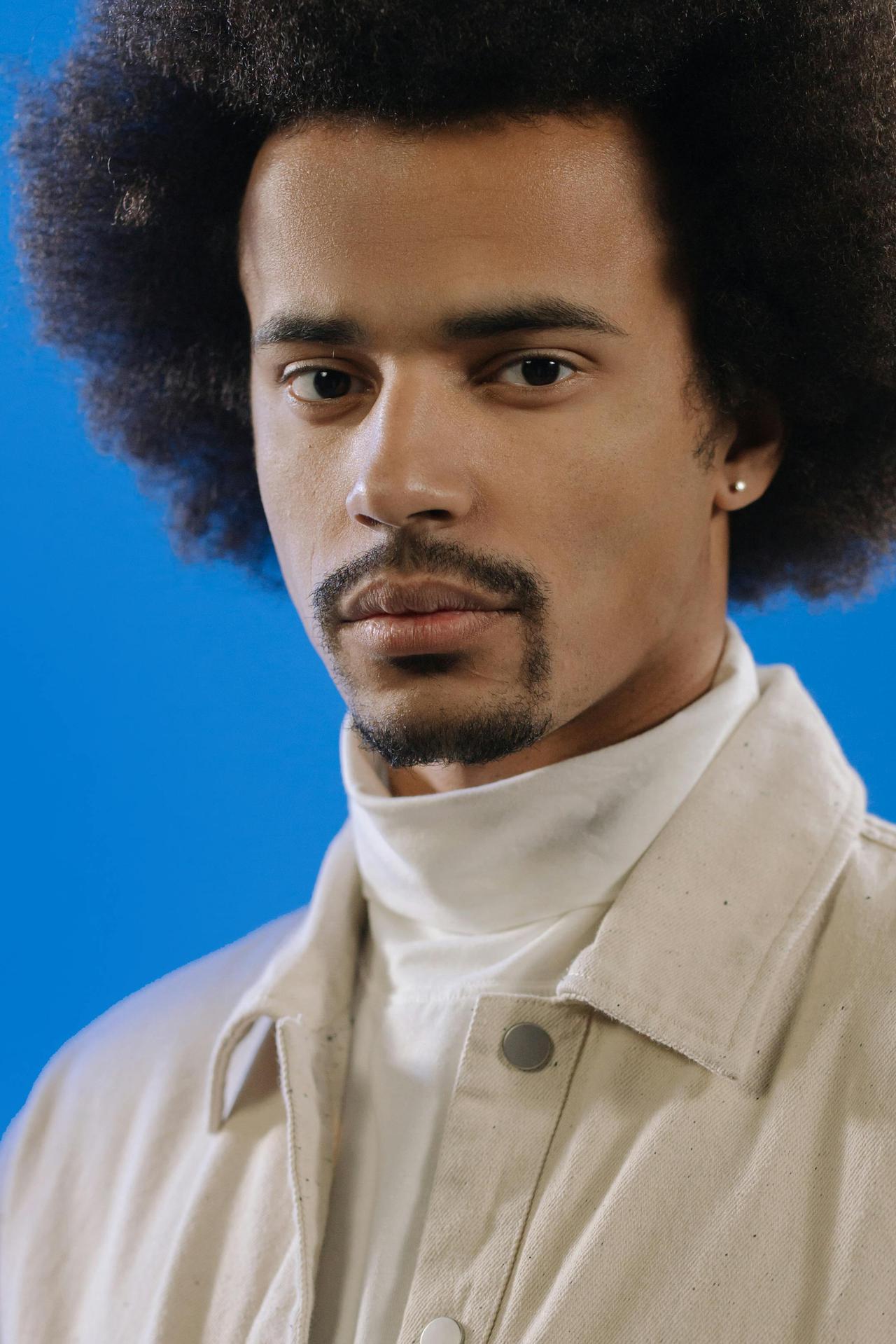Studio portrait of a man with an afro hairstyle and goatee against a blue backdrop, wearing a white jacket.