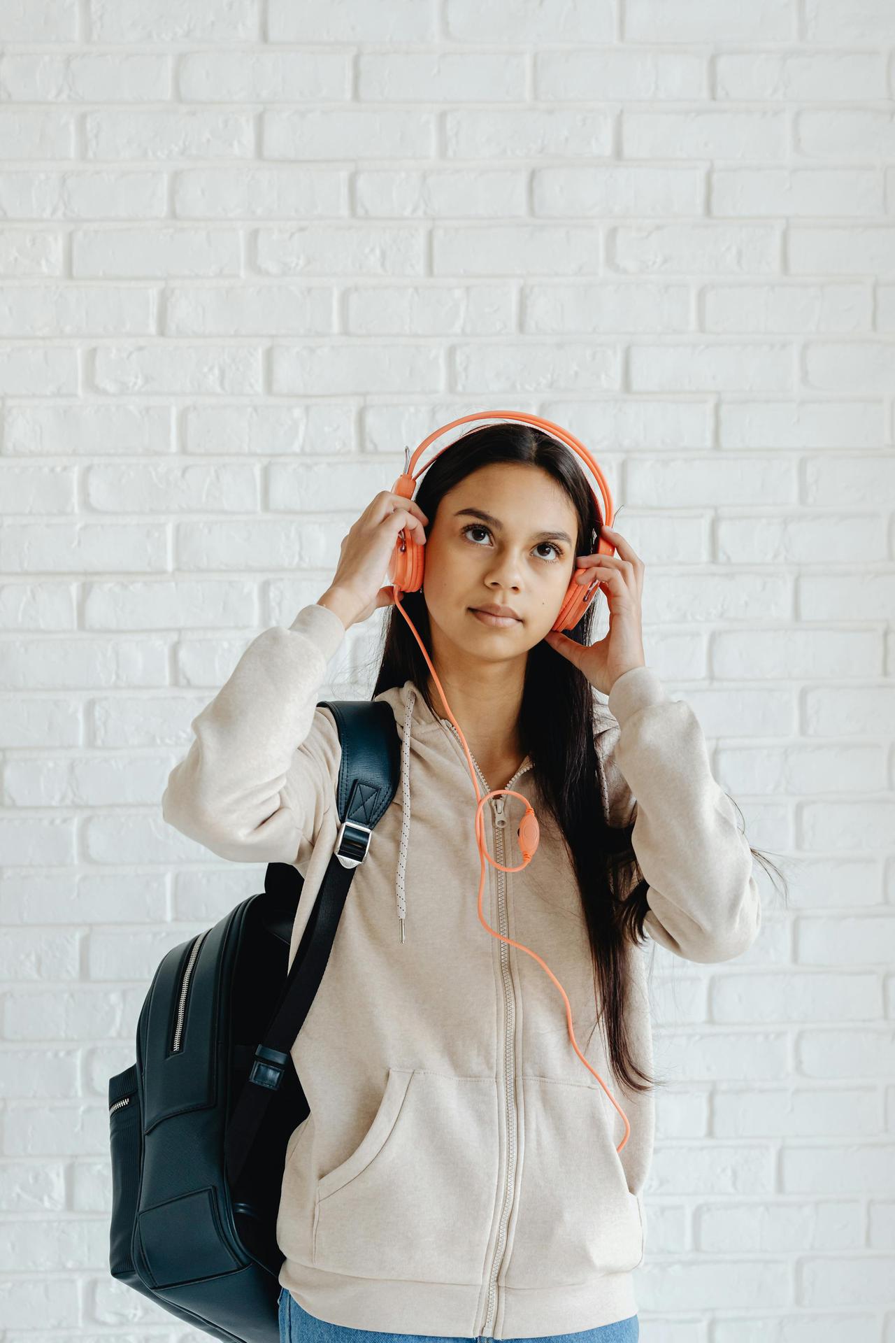 Teenage girl wearing headphones and backpack against white brick wall.