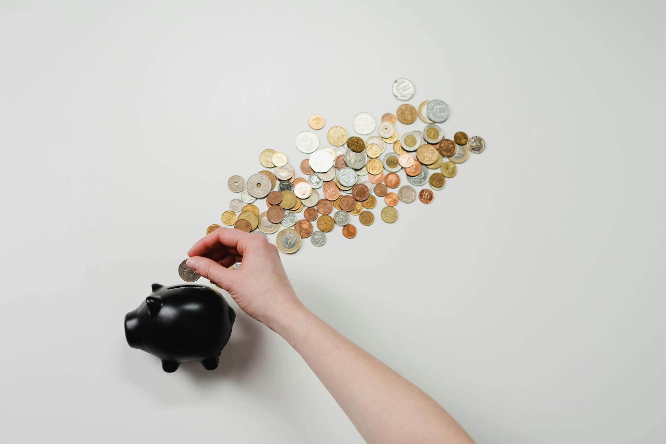 A person placing a coin into a black piggy bank with scattered coins around it, symbolizing how financial factors influence therapy prices and affordability at Cognitive Analytica in Lebanon.